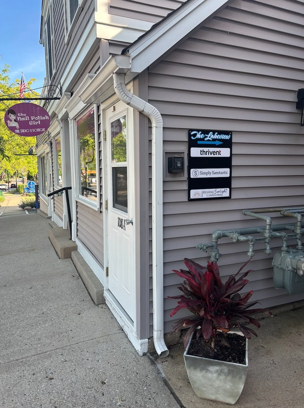 Side view of a commercial building with signs indicating it houses businesses like The Nail Polish Girl, Thrive, Simply Samtastic, and Rising Sunlight Counseling. There is a potted red-leaf plant outside, and a sidewalk with a street in the backgroun