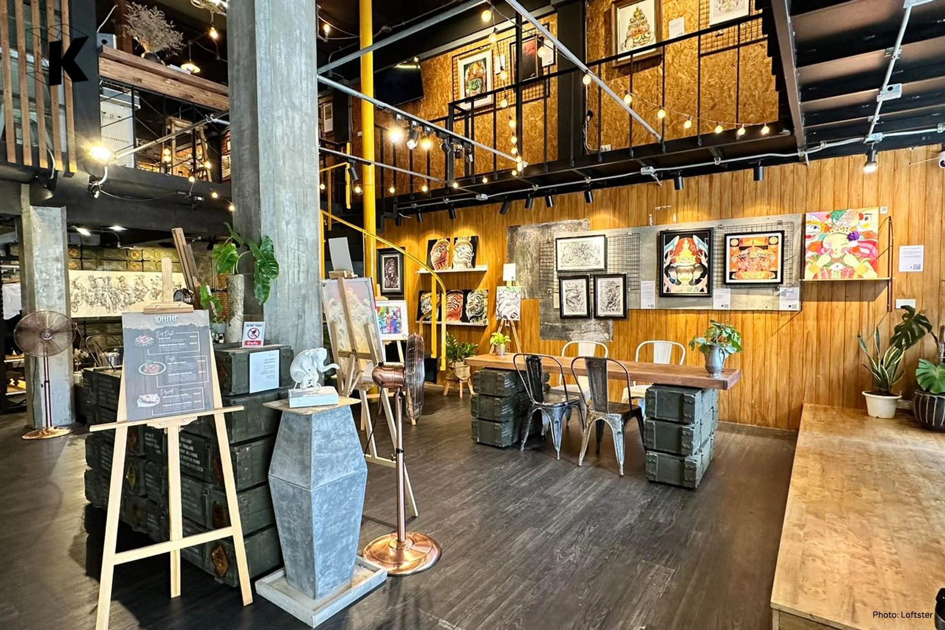 Interior view of an art gallery or cafe with wooden walls, framed artwork displayed, and a table with chairs and potted plants.