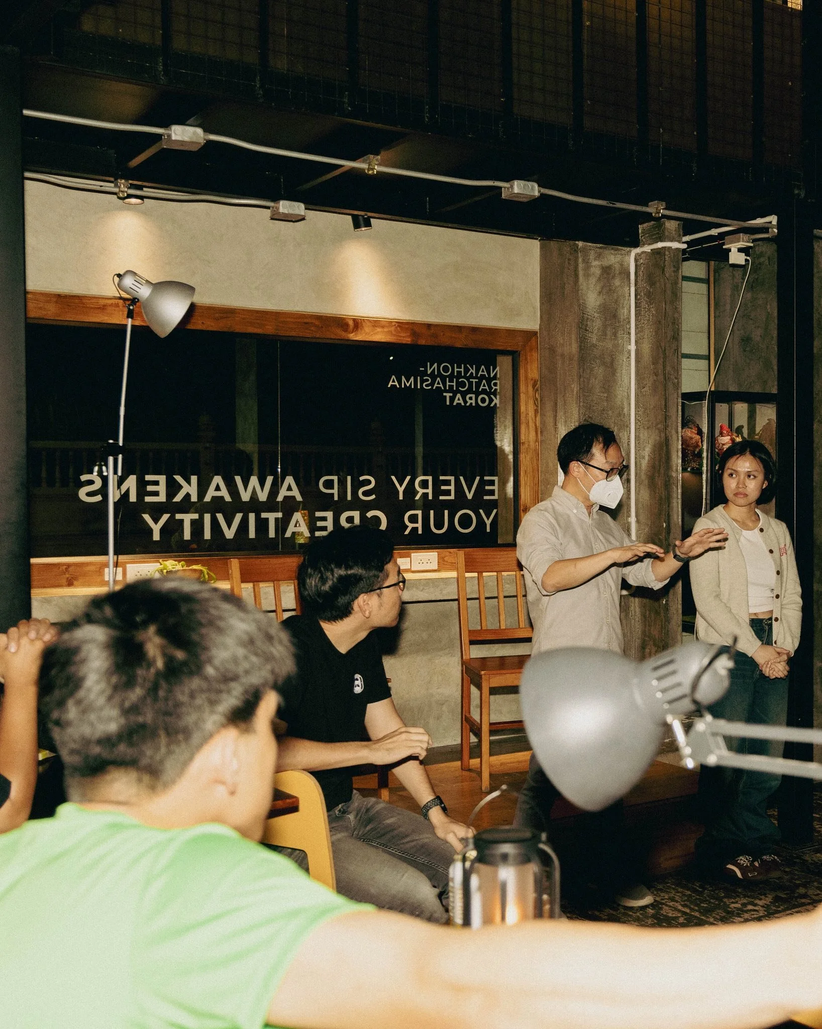 A group of people attending a presentation or discussion in a café or restaurant with a sign that reads 'Every sip awakens your creativity.' One man, wearing a face mask, is speaking to the group while two women and a man listen.