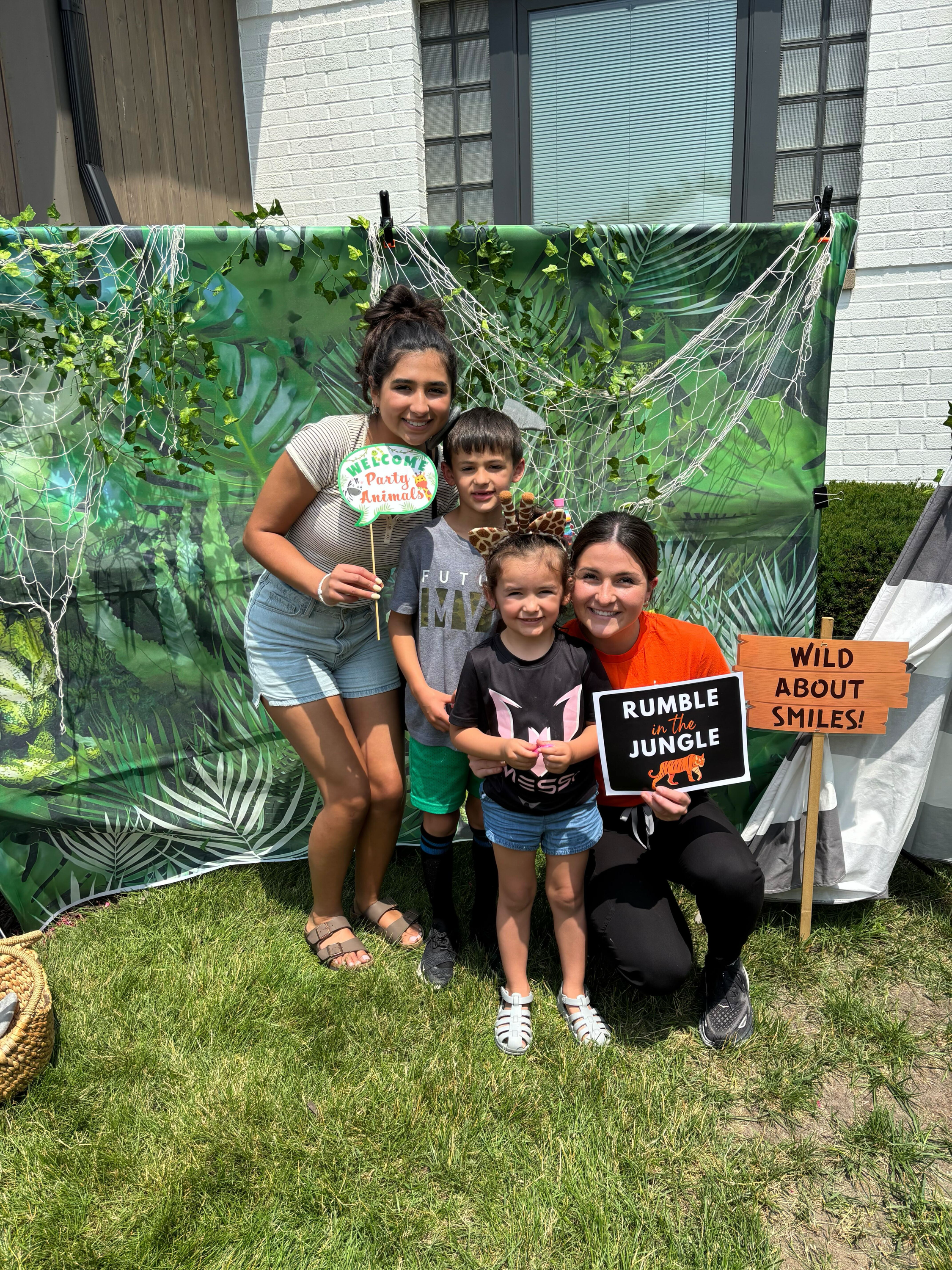 Group of four people, two adults and two children, at a jungle-themed photo booth with green leafy background and spider web decorations. The children are holding animal-themed props, and one of the adults is holding a sign that says 'Welcome Party A