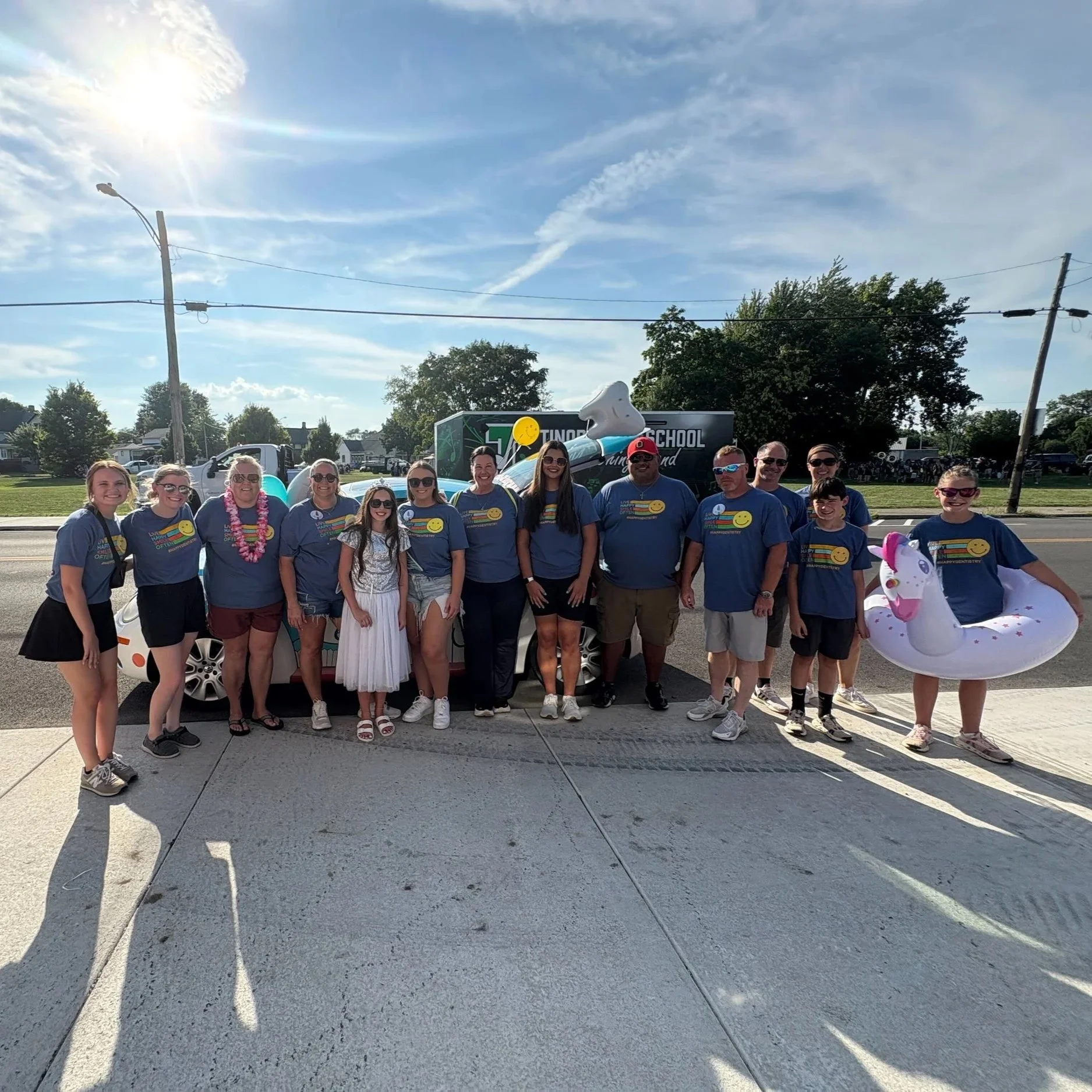 Group of people wearing blue shirts standing in front of a colorful car with a unicorn float and a large paddle toy, on a sunny day.