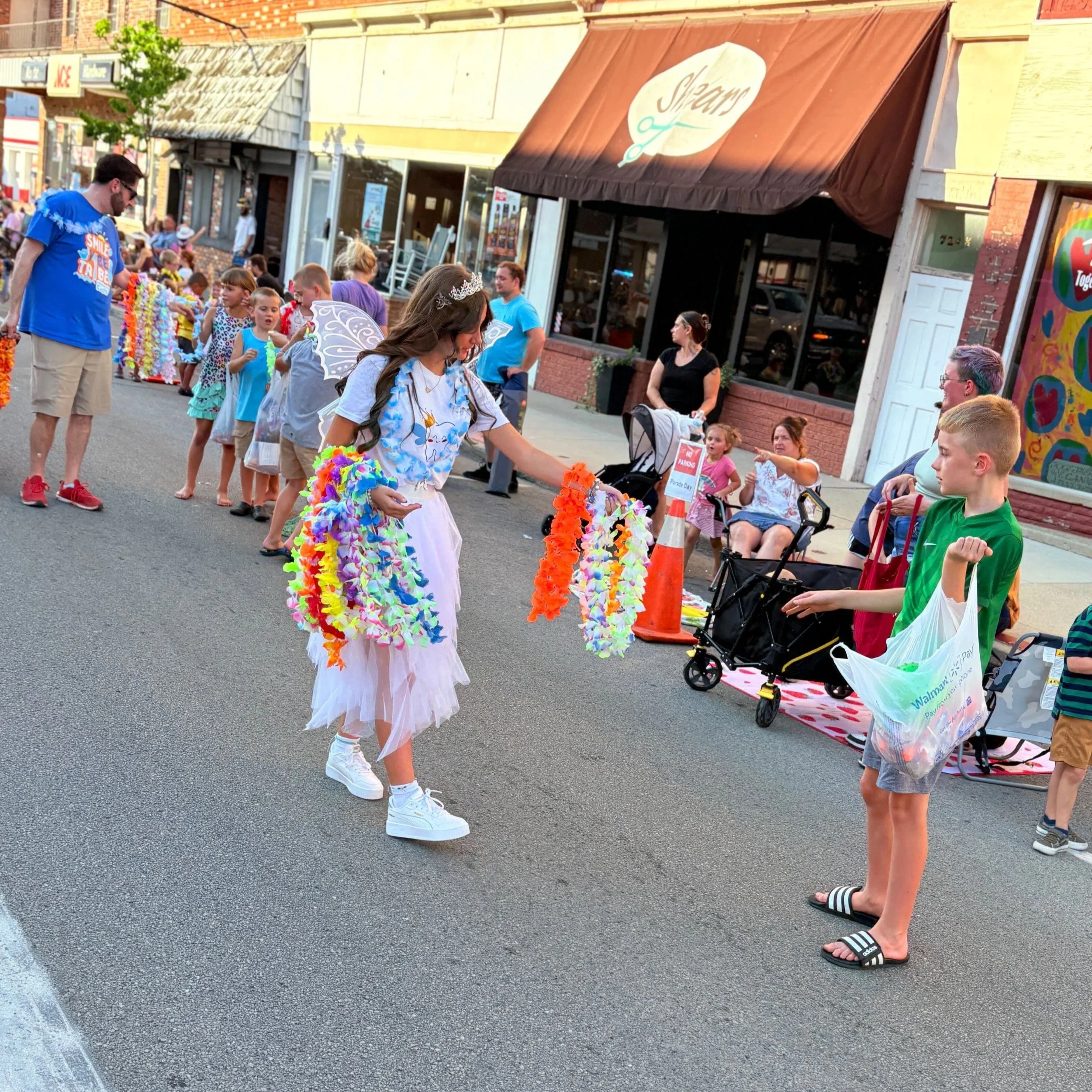 A young girl dressed as a fairy with wings and a tiara is holding colorful leis in a street parade. She is standing in front of a boy wearing a green shirt and sandals, holding a shopping bag. People are sitting and standing along the sidewalk, watch