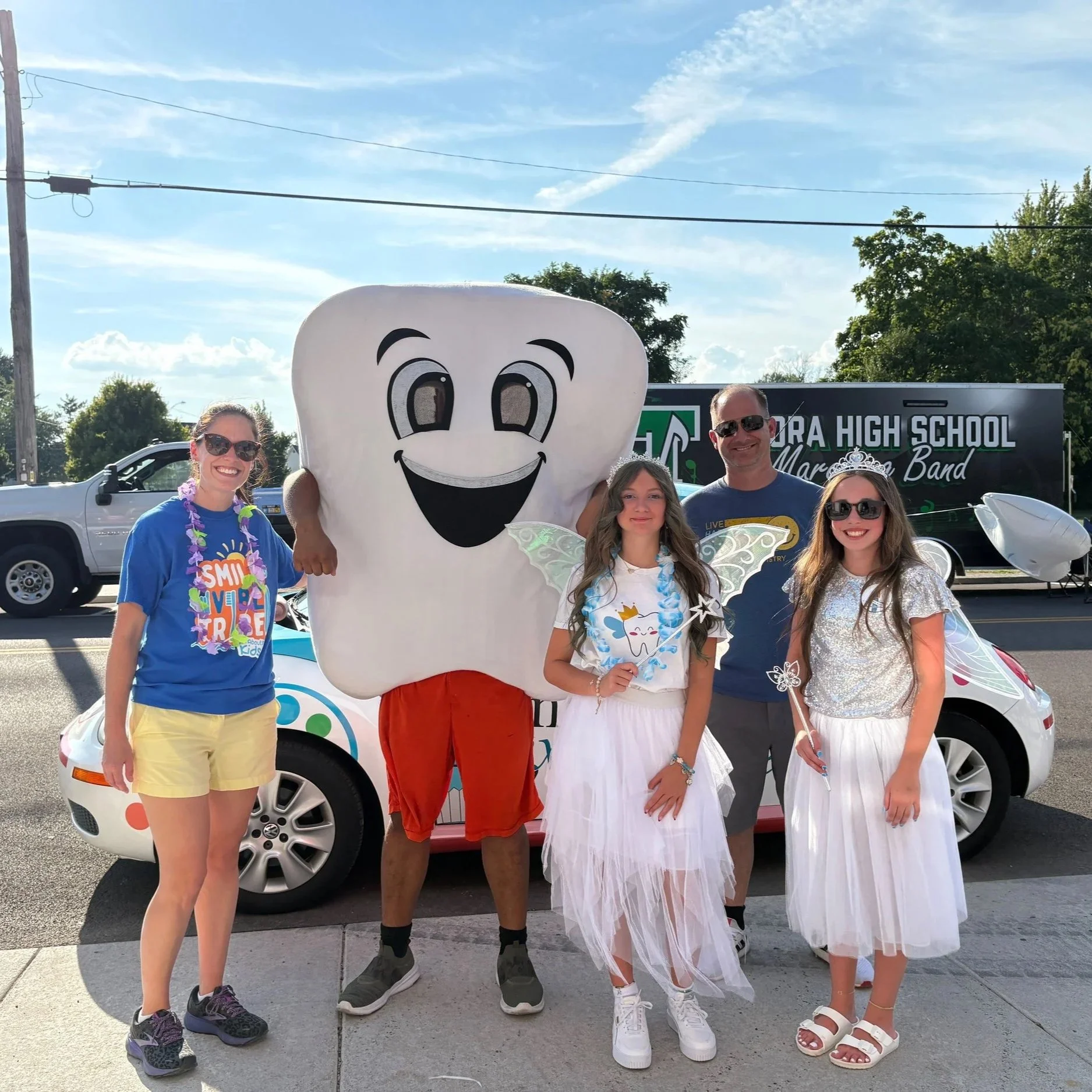 Group photo of five people, including a person in a large cartoon tooth costume, standing outdoors on a sunny day, in front of a high school band truck, with two girls dressed as fairies and a woman in a blue shirt and yellow shorts, all smiling.