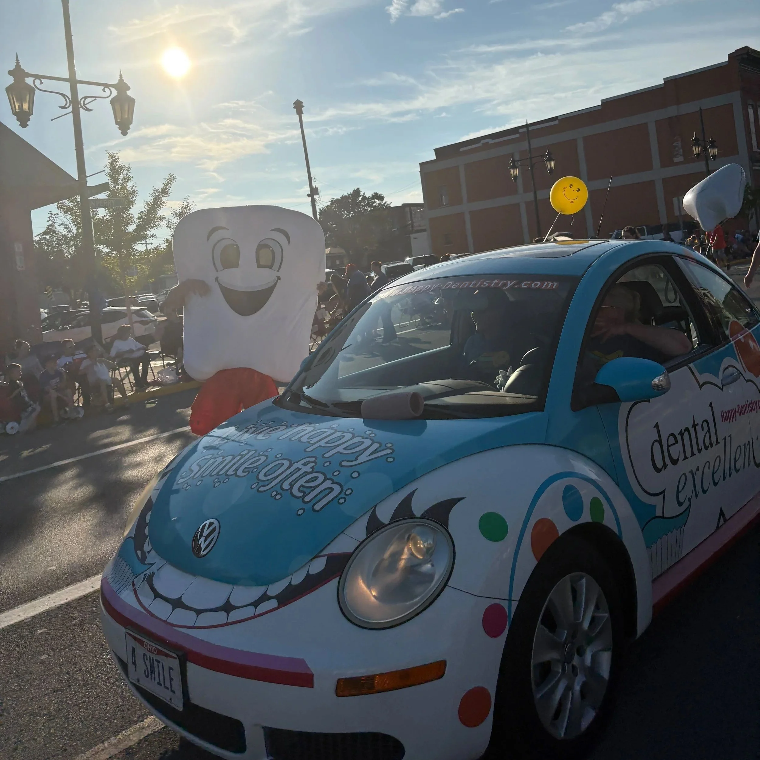 A decorated Volkswagen Beetle with a smiling teeth design, parked on a street during a parade or event. There is a large inflatable cartoon tooth and a yellow smiley face balloon on top of the car. People and buildings are visible in the background.