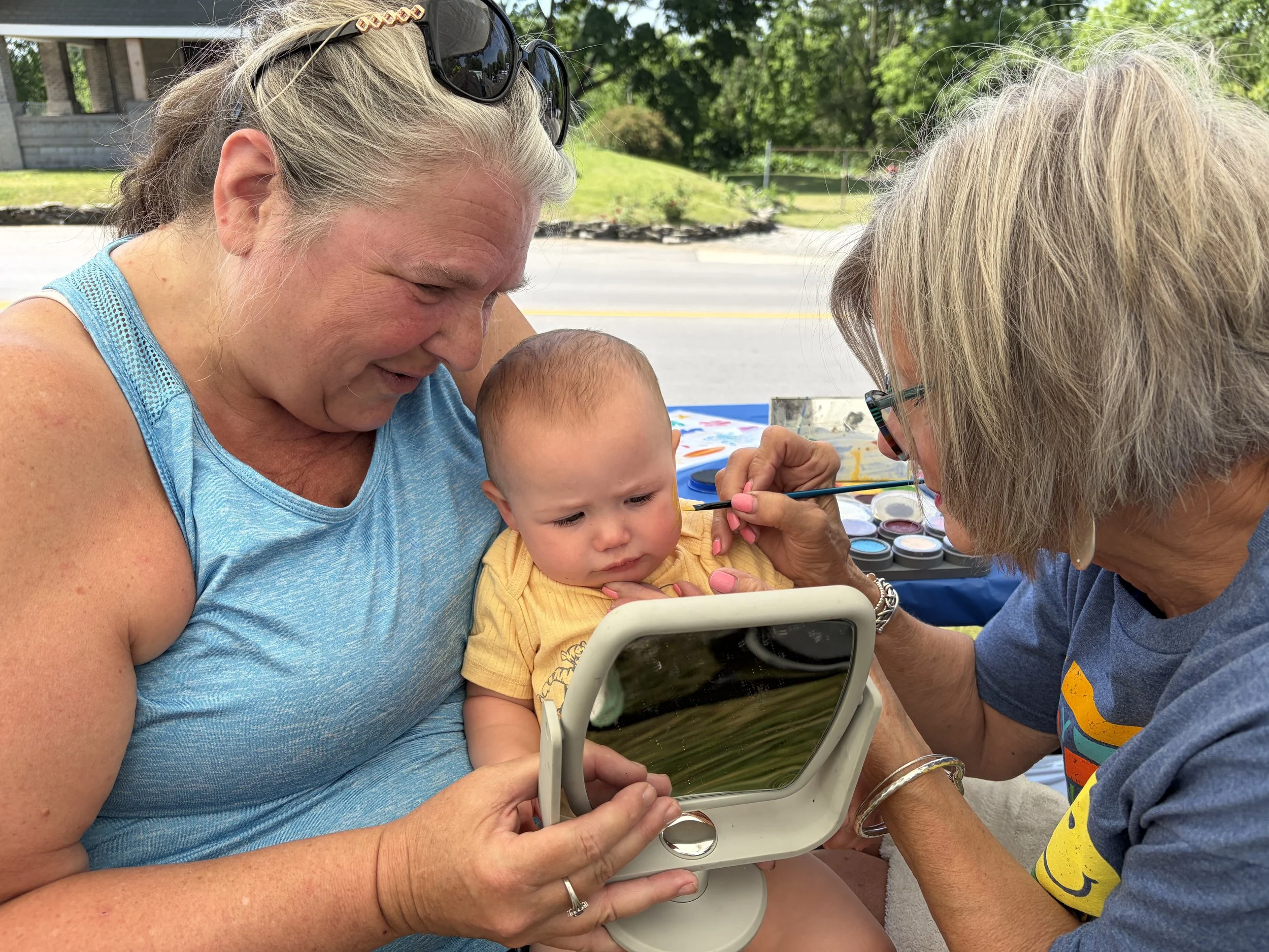 A woman holding a young child with a third woman painting the child's face with a brush. They are outdoors on a sunny day near a road and green trees.