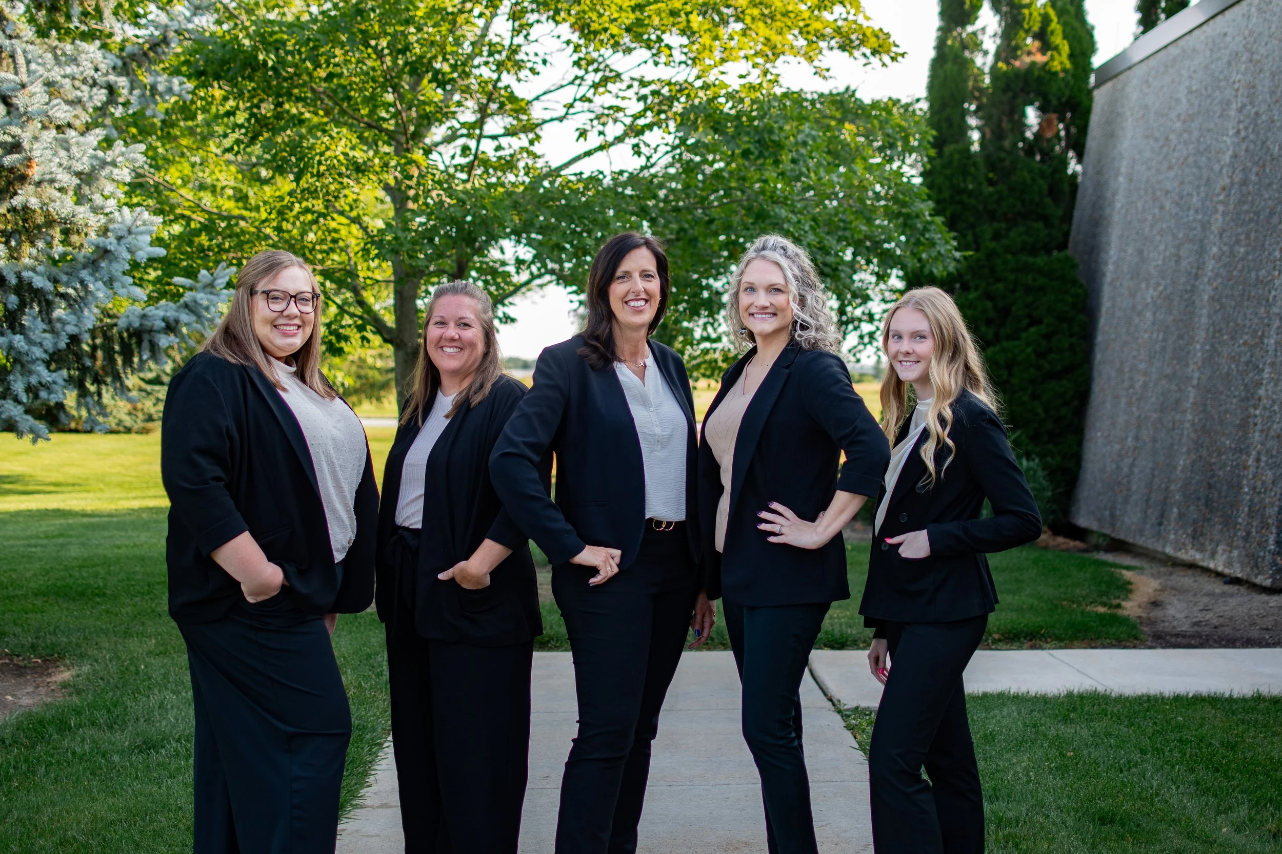 Group of five women standing outdoors on a sidewalk, smiling, with trees and a stone wall in the background.