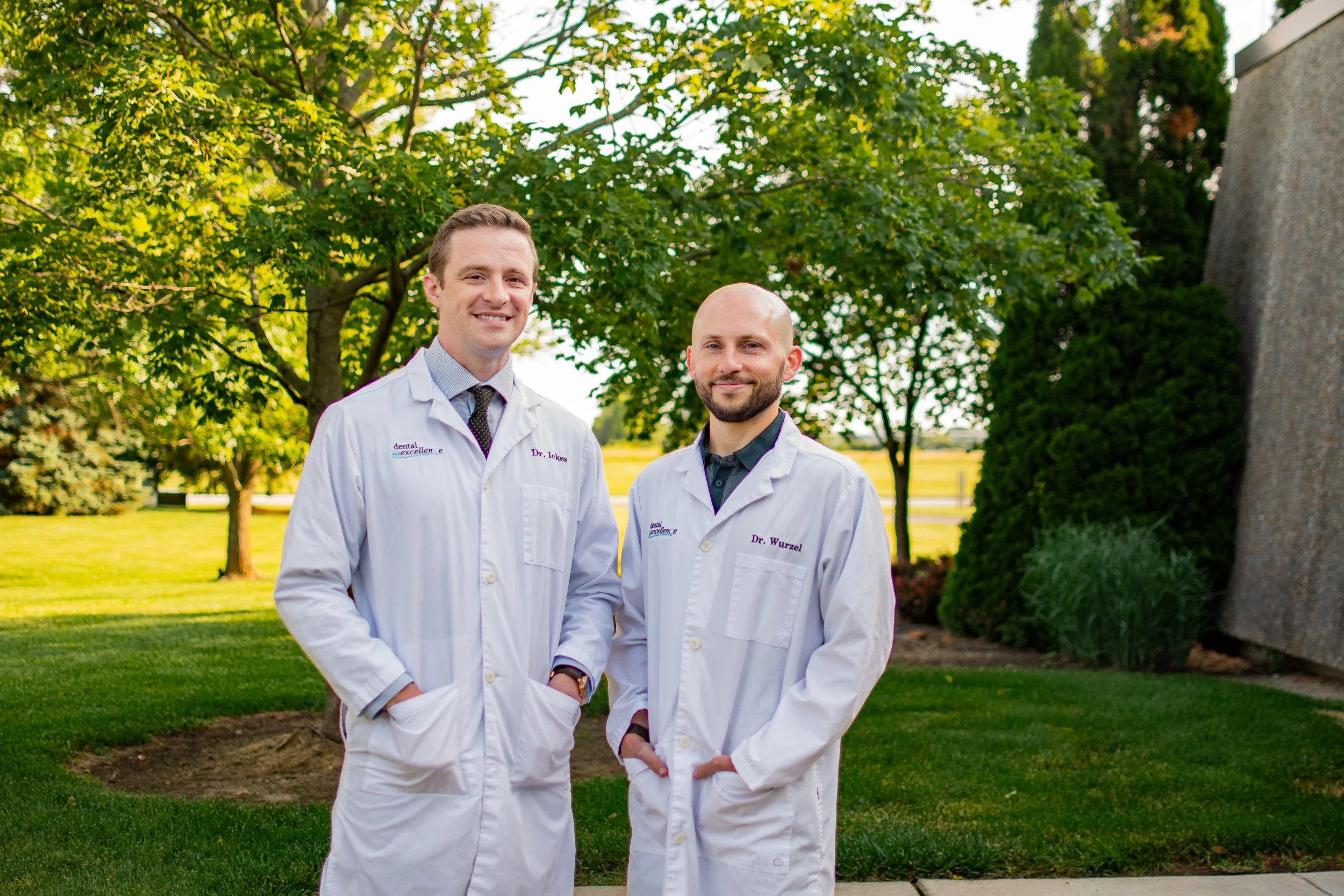 Two male dentists standing outdoors in a park. One is smiling, wearing a white coat with a name badge that reads Dr. Ickes. The other is also smiling, with a white coat and badge reading Dr. Wurzel. They are surrounded by lush green trees and grass.