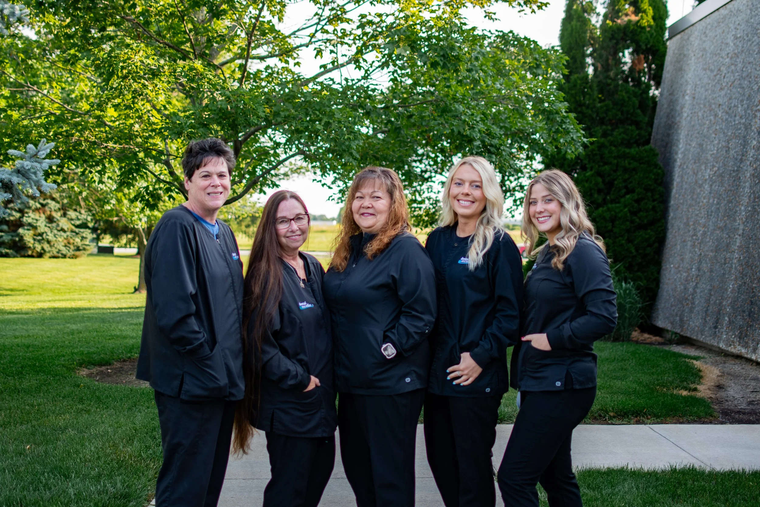 Group of five women standing outdoors on a grassy area, smiling, with trees and a building in the background.