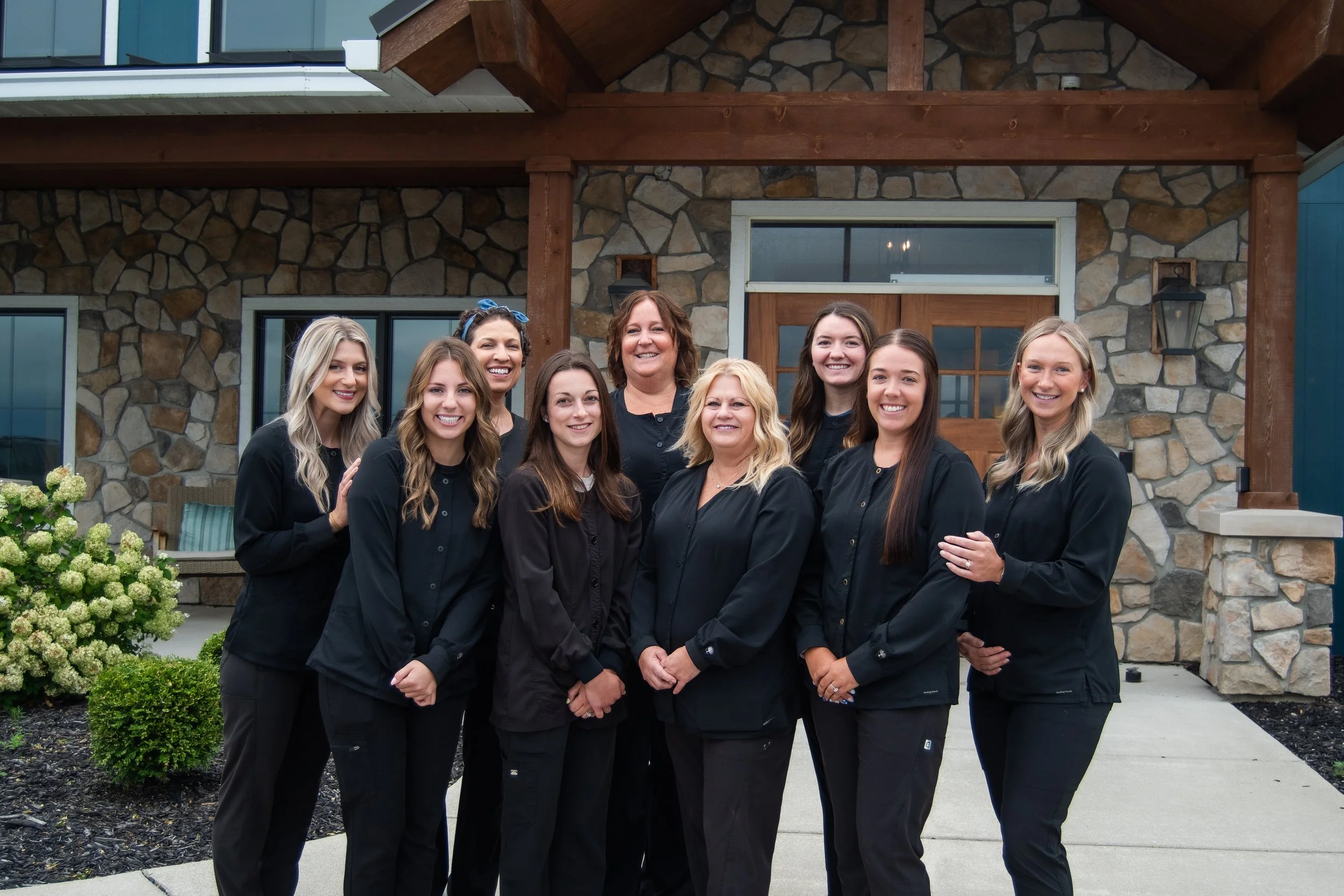 Group of nine women standing outside in front of a stone and wood house, smiling at the camera.