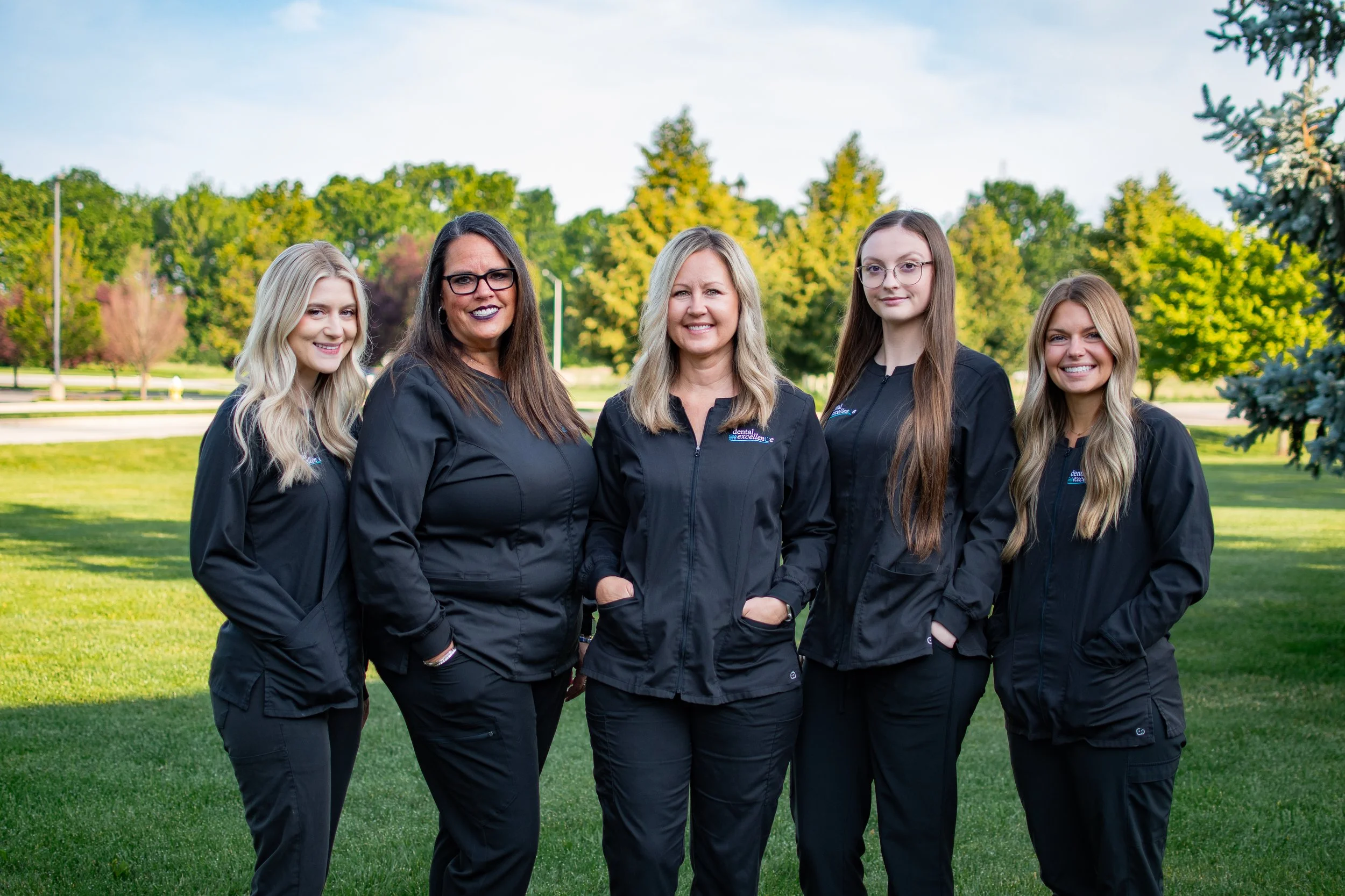 Group of five women in black jackets standing outdoors in a park with green trees and grass.