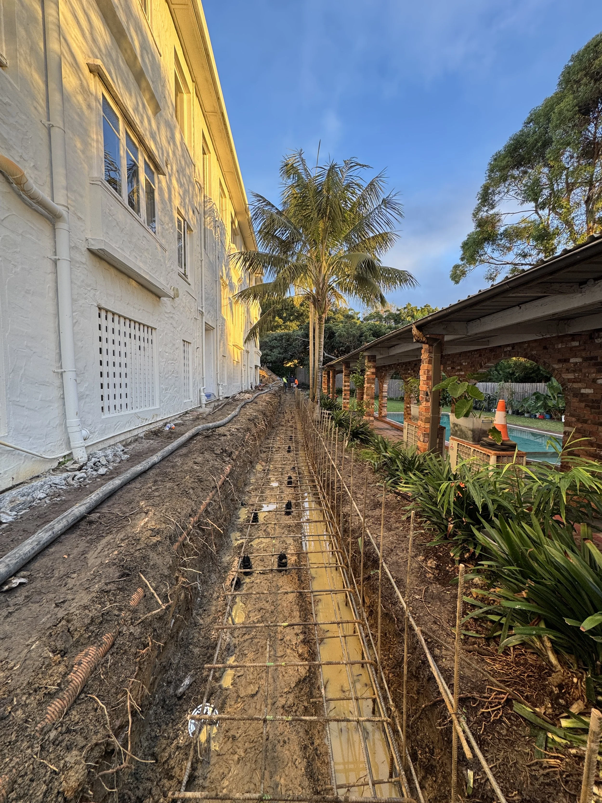 Construction site with exposed rebar and pipes, dirt trench alongside a white stucco building and brick pergola, with trees and a pool in the background, during sunset.