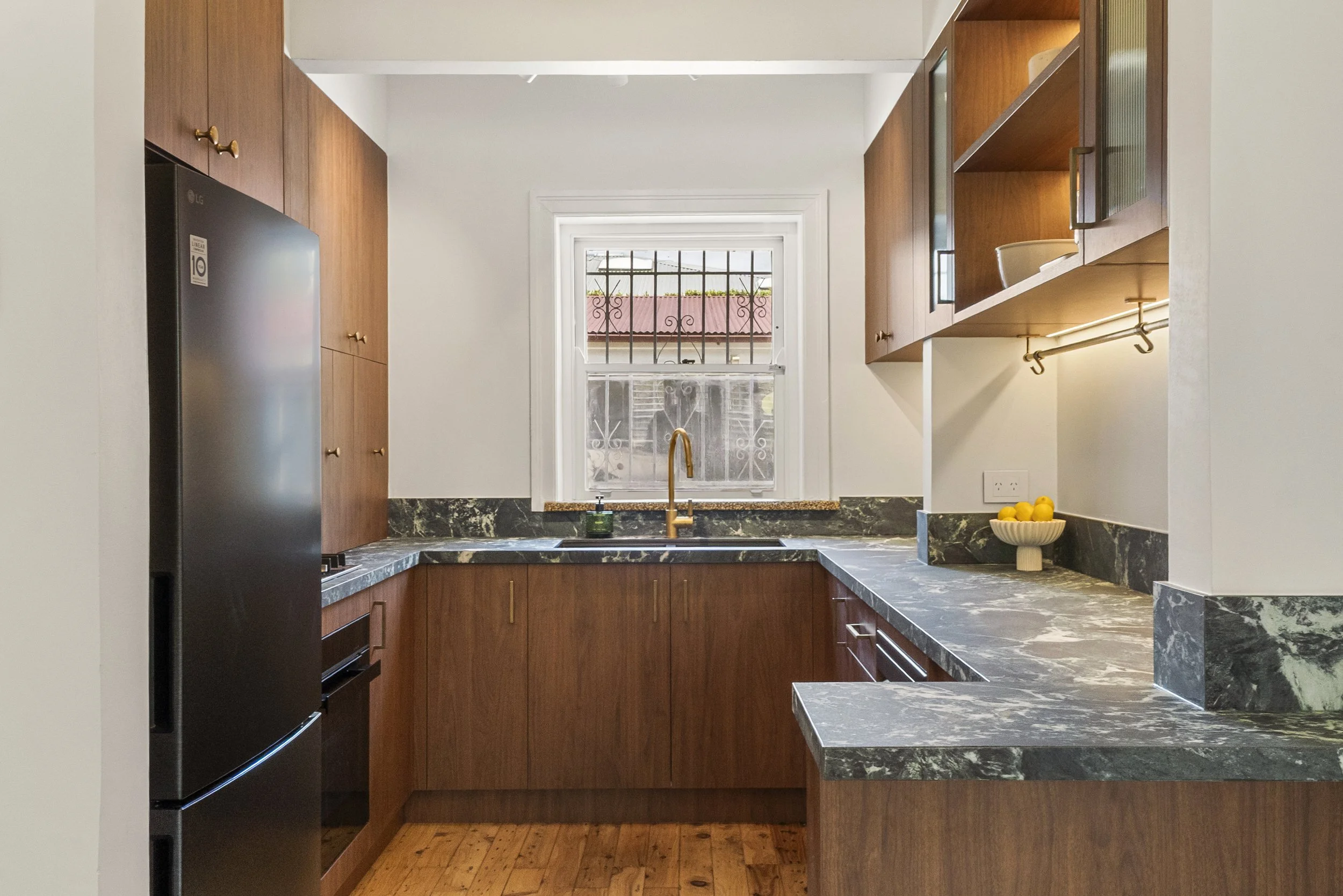 A kitchen with wooden cabinets, a black refrigerator, and marble countertops, located in front of a window with a barred guard inside. There are yellow lemons in a bowl on the counter.
