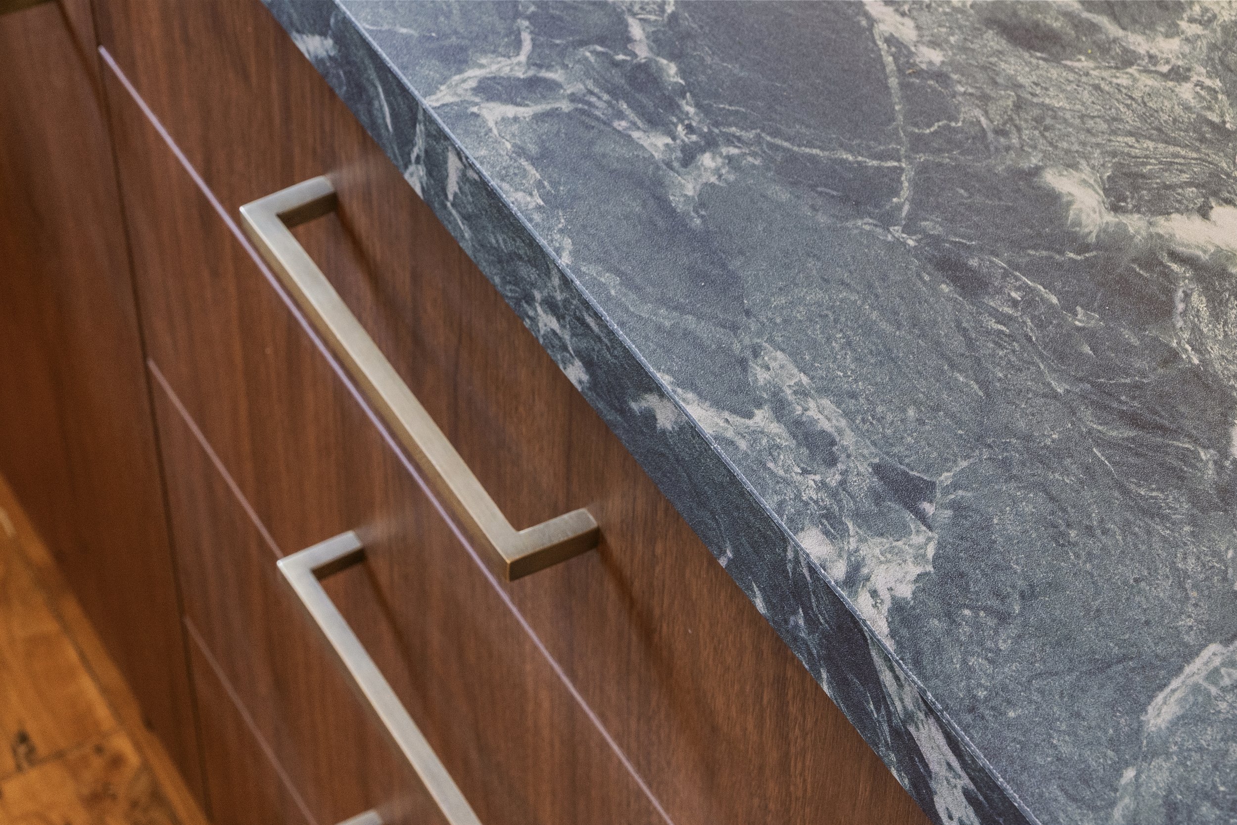 Close-up view of a dark gray marble countertop with white veining, part of a kitchen or bathroom setup, with a wooden cabinet with horizontal drawer handles underneath.