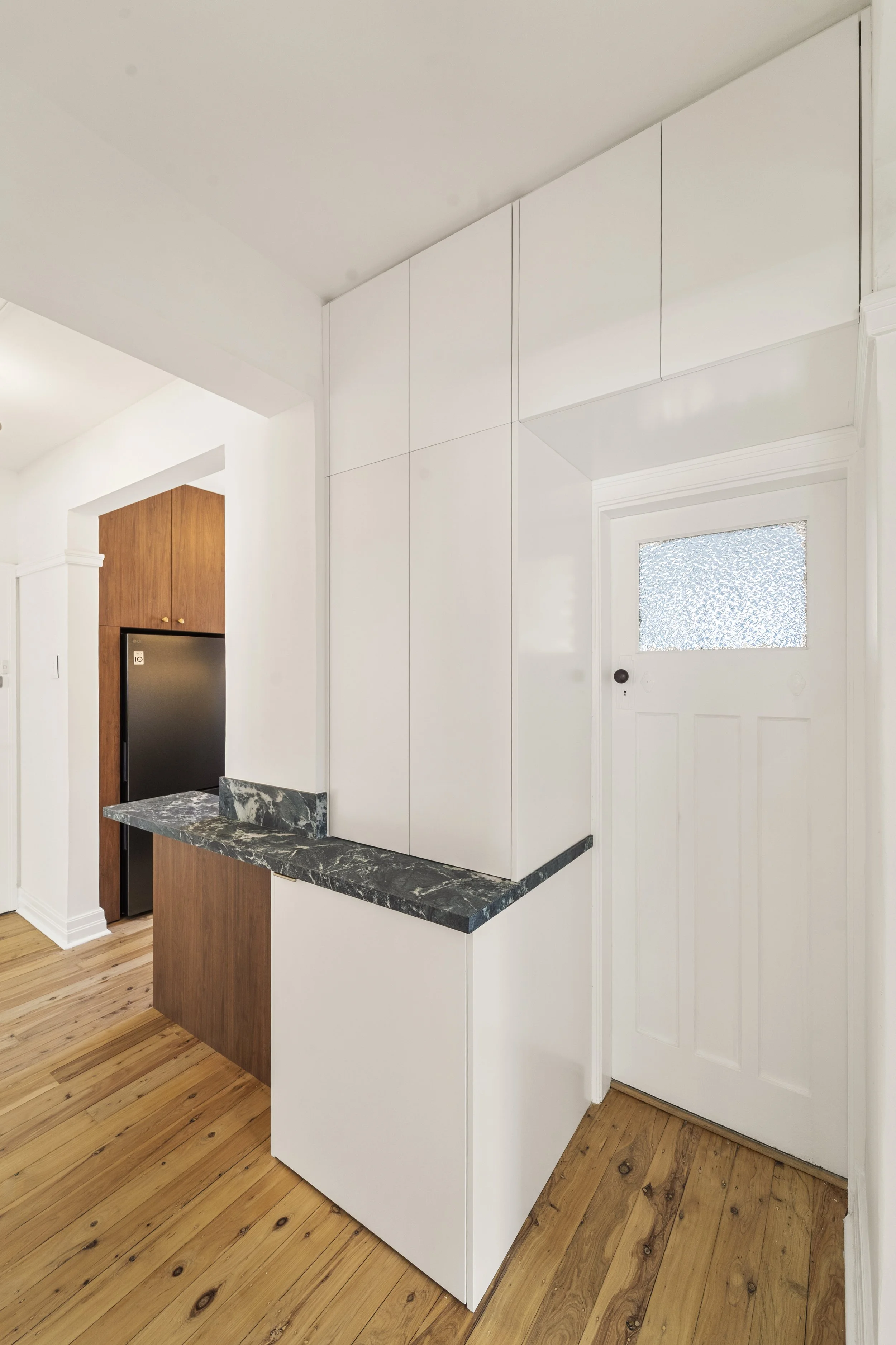 Interior of a modern kitchen with white cabinets, a black granite countertop, and a white door with frosted glass, next to a wooden floor.