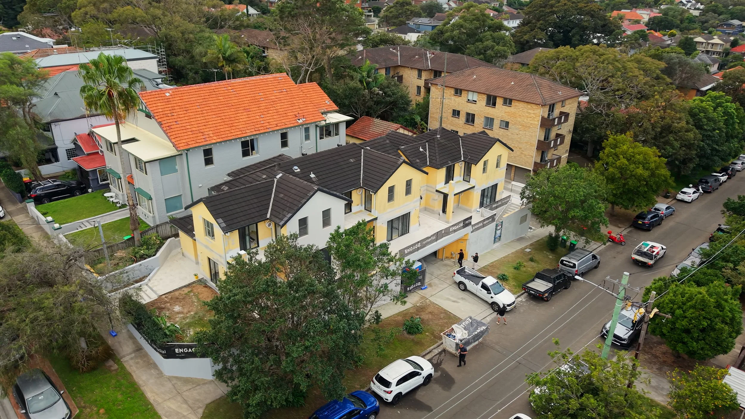 Aerial view of a neighborhood with various houses and apartment buildings, parked cars, trees, and a street with people walking. There are multi-story residential buildings, some with red and dark-colored roofs, and a mix of lush green trees.