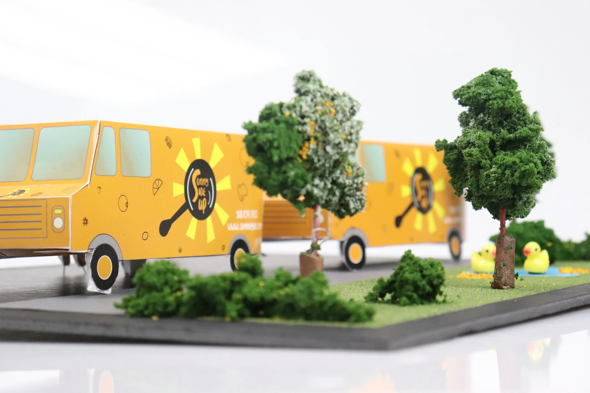 Miniature food trucks with a yellow design, trees, bushes, and rubber ducks on a model landscape.