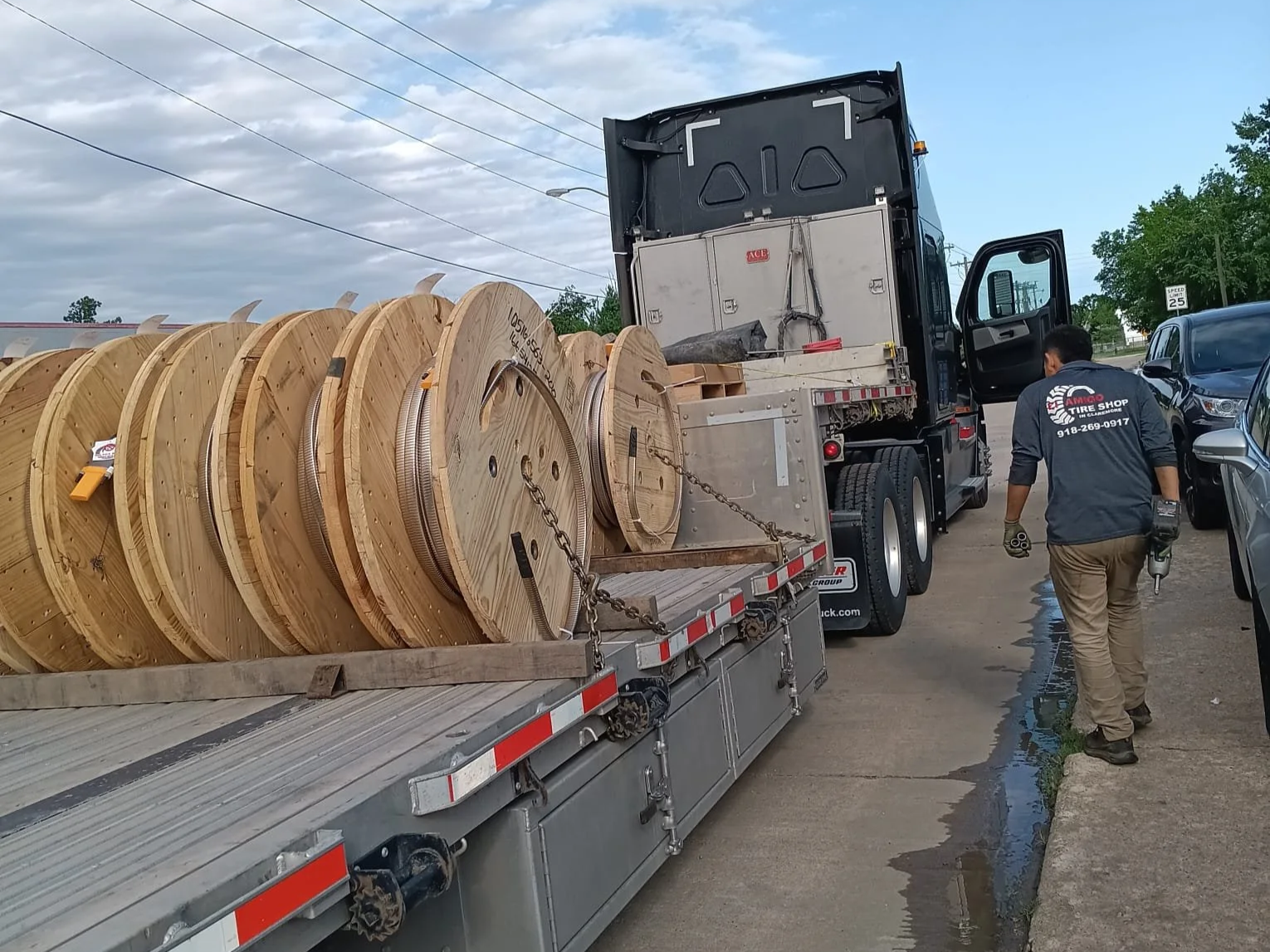 A man walking beside a truck loaded with large wooden spools on a street with parked cars and a 25 mph speed limit sign.