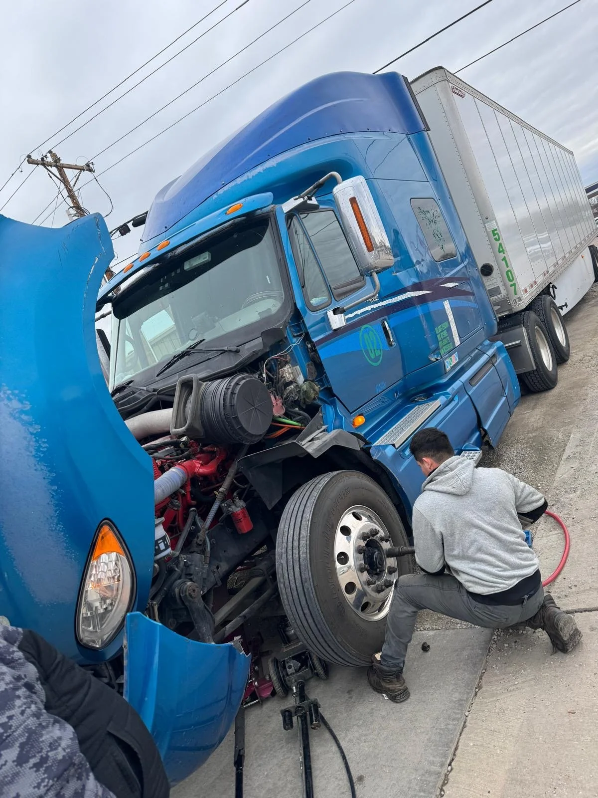 A person working on a blue semi-truck that appears to have a flat tire or mechanical issue, on a concrete surface under an overcast sky.