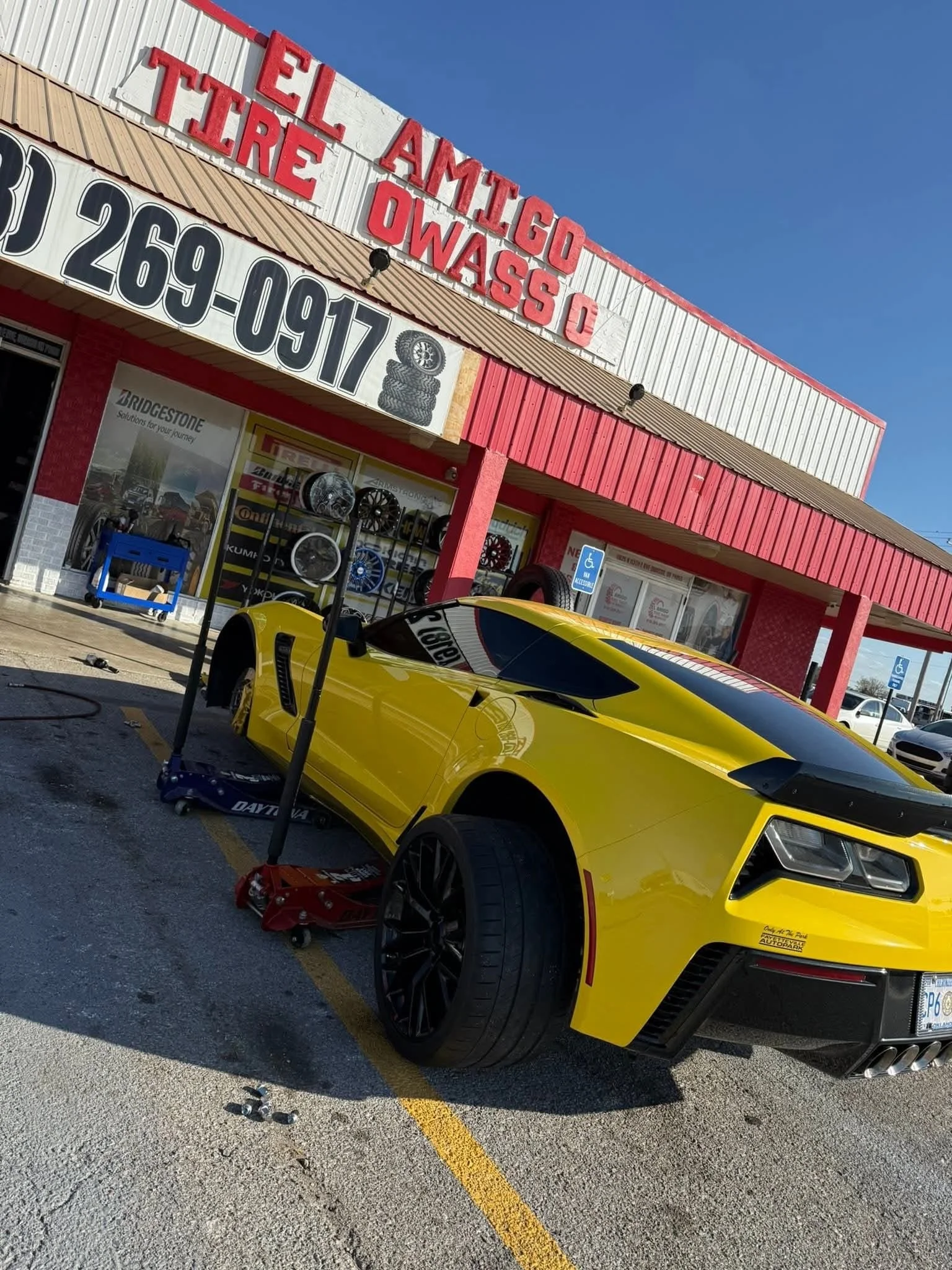 A yellow sports car being repaired outside an auto shop named 'El Amigo Tire O.' The car is on a jack, with tools and equipment nearby, in a parking lot with handicapped parking spaces.