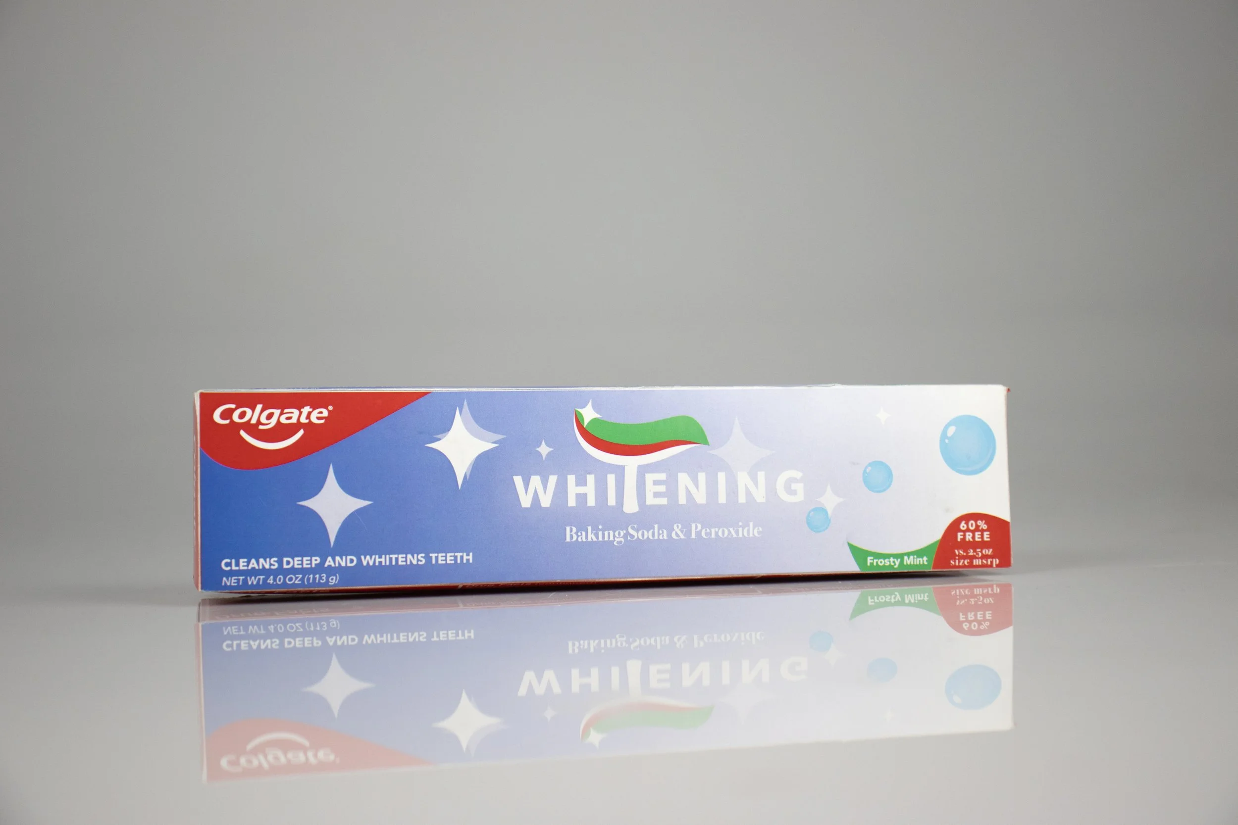 A box of Colgate Whitening Baking Soda & Peroxide toothpaste with frosty mint flavor, featuring blue packaging, white stars, and a red and green logo, reflected on a glossy surface.