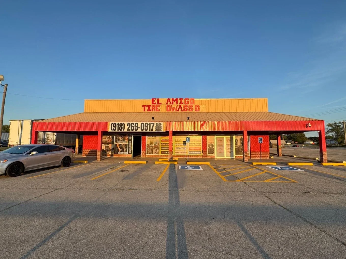 A tire shop called 'El Amigo Tire Owaso' with a yellow and red exterior, parked cars, and marked handicap parking spots in front, during daytime with a clear sky.