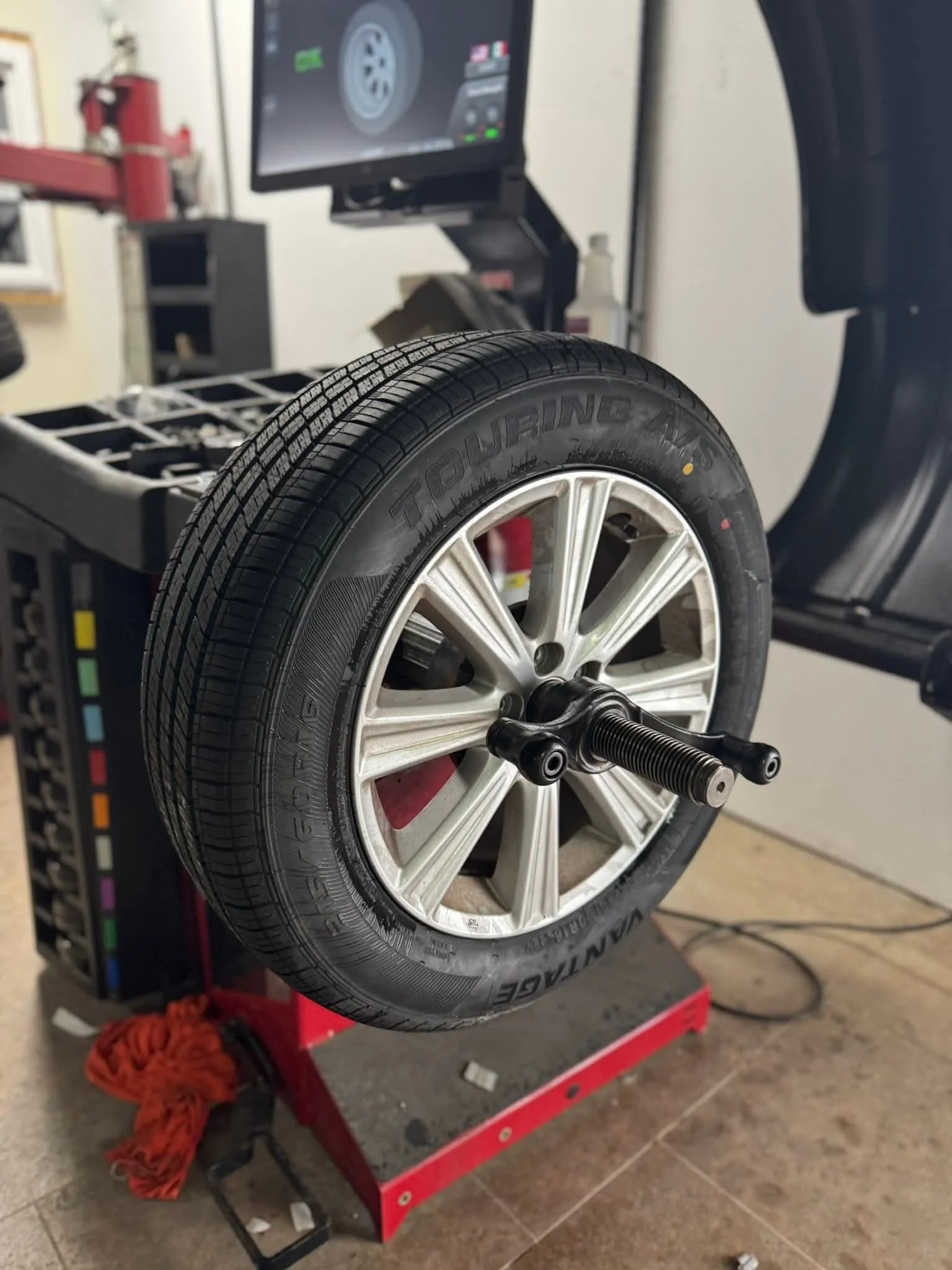 A car wheel mounted on a testing machine with a tire labeled 'Touring A/S' and a digital monitor in the background displaying wheel data.