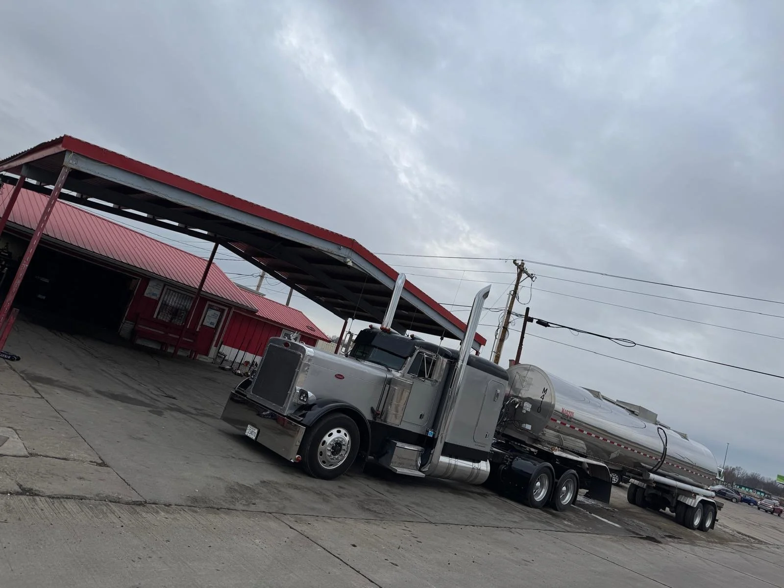 A silver tanker truck parked in a lot under an overcast sky with utility poles and a red building in the background.