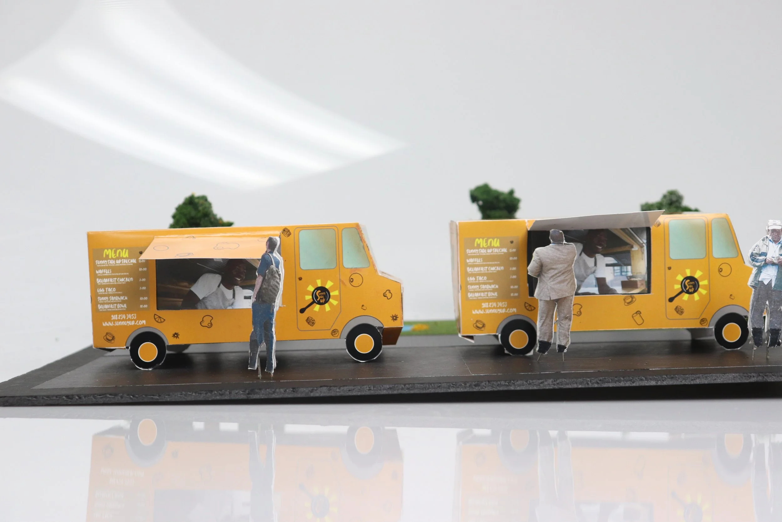 Miniature yellow food trucks with printed menus, along with paper cutout figures of people, displayed on a black platform.
