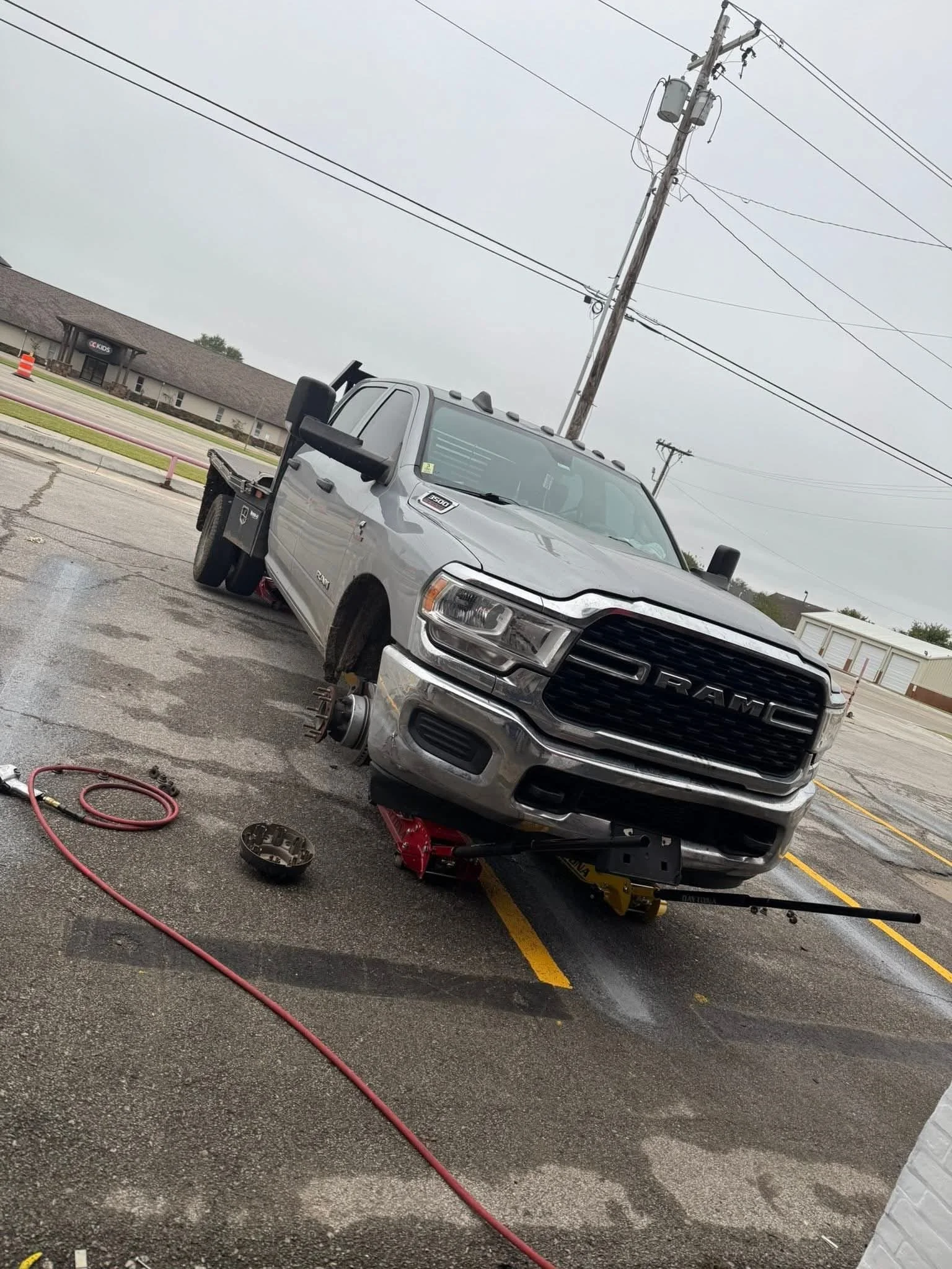 A silver RAM pickup truck is lifted on a hydraulic jack with one front wheel removed in a parking lot on a cloudy day. Various tools and parts are scattered on the ground.