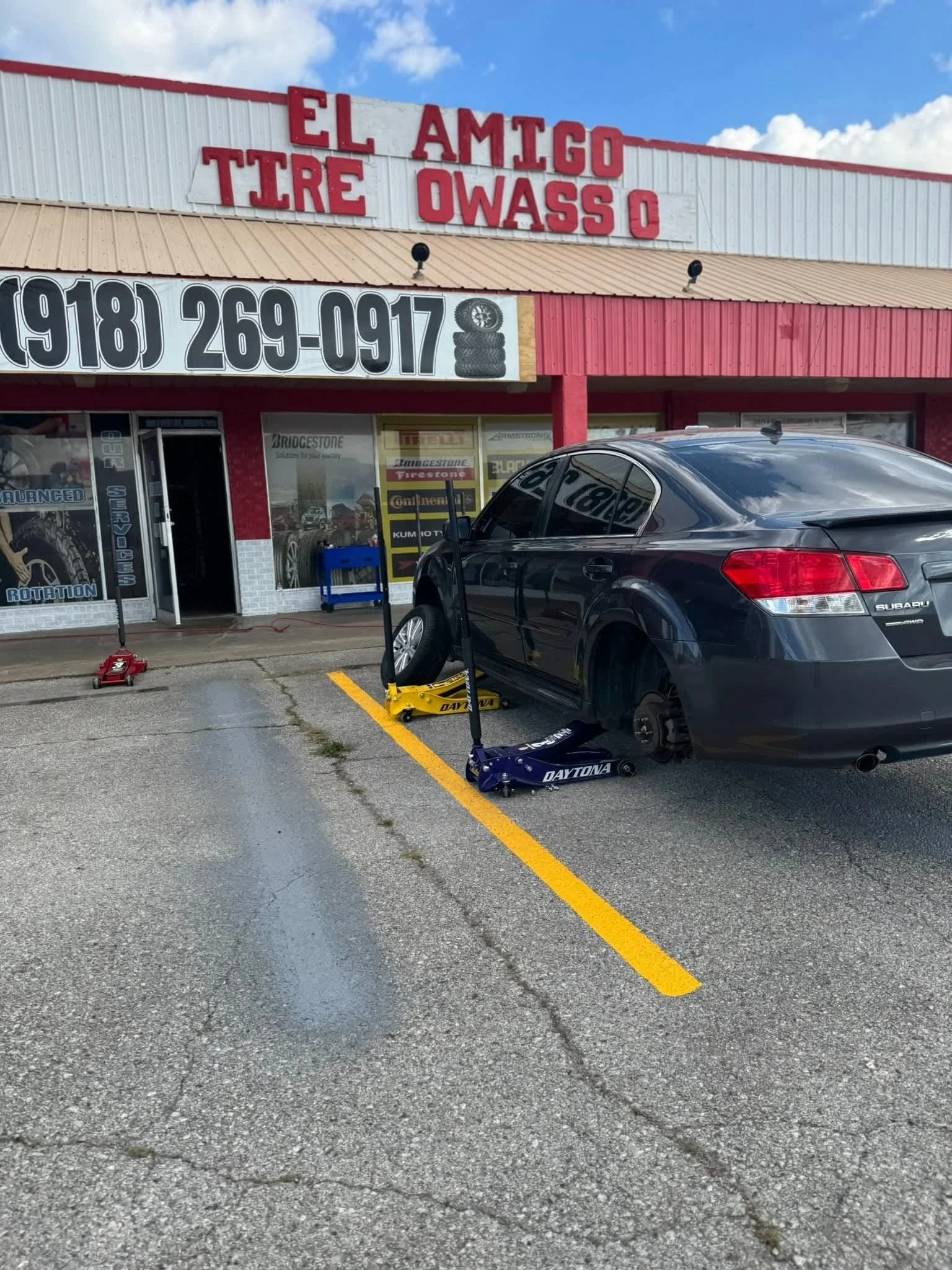 A black car elevated on jack stands outside of an auto repair shop with a sign reading 'El Amigo Tire & Oass' and a large phone number. The shop has red and white exterior walls, and the car appears to be undergoing tire or wheel service.