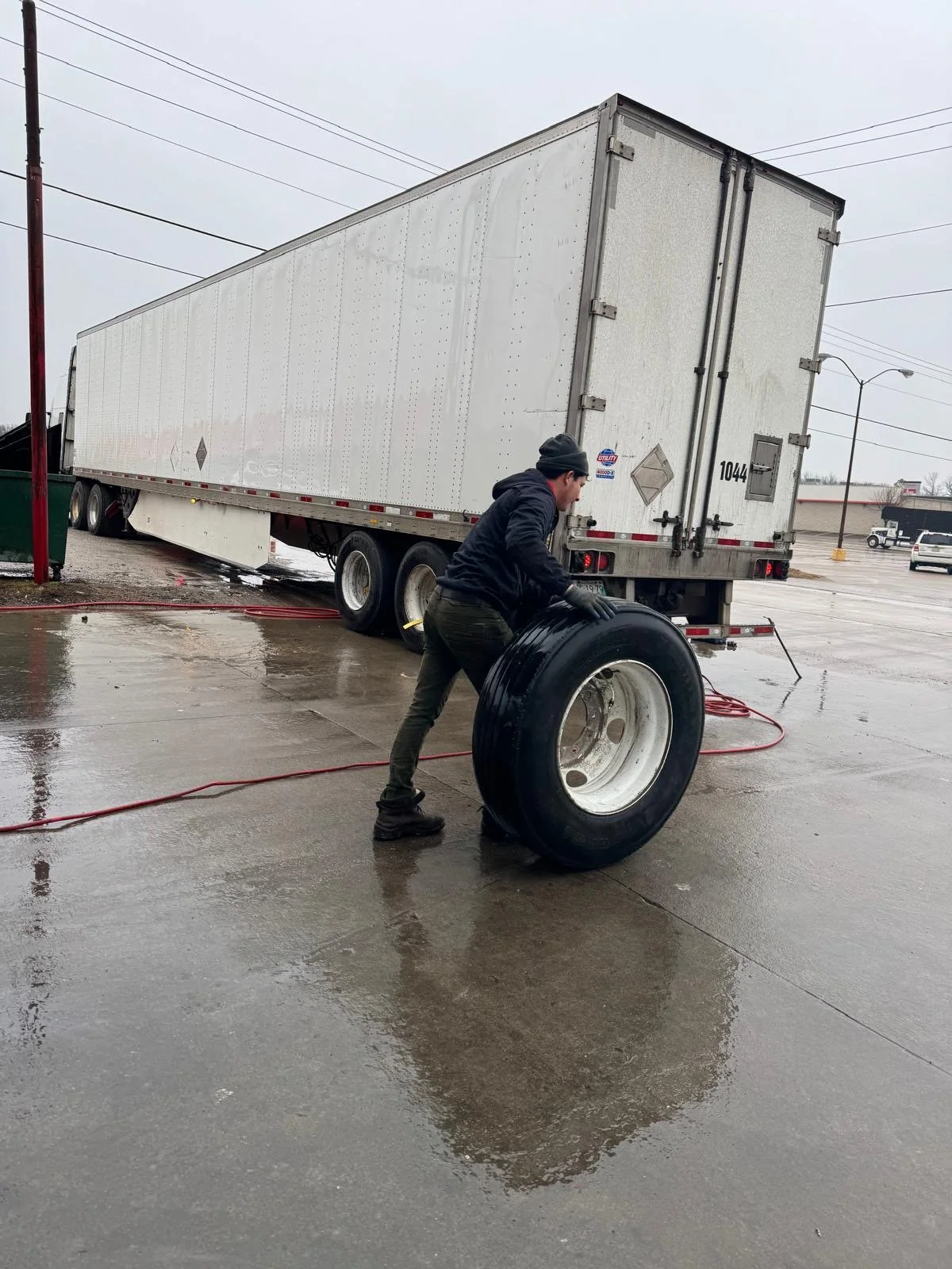A person is changing a tire on a wet concrete surface near a large white semi-truck trailer on an overcast day.