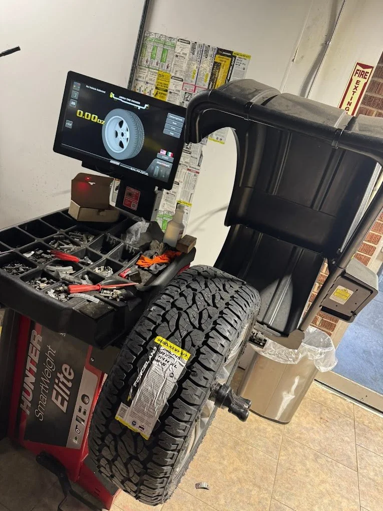 Automotive tire balancing machine with a large tire mounted on it, and a computer screen displaying wheel balancing data, located inside a service station.