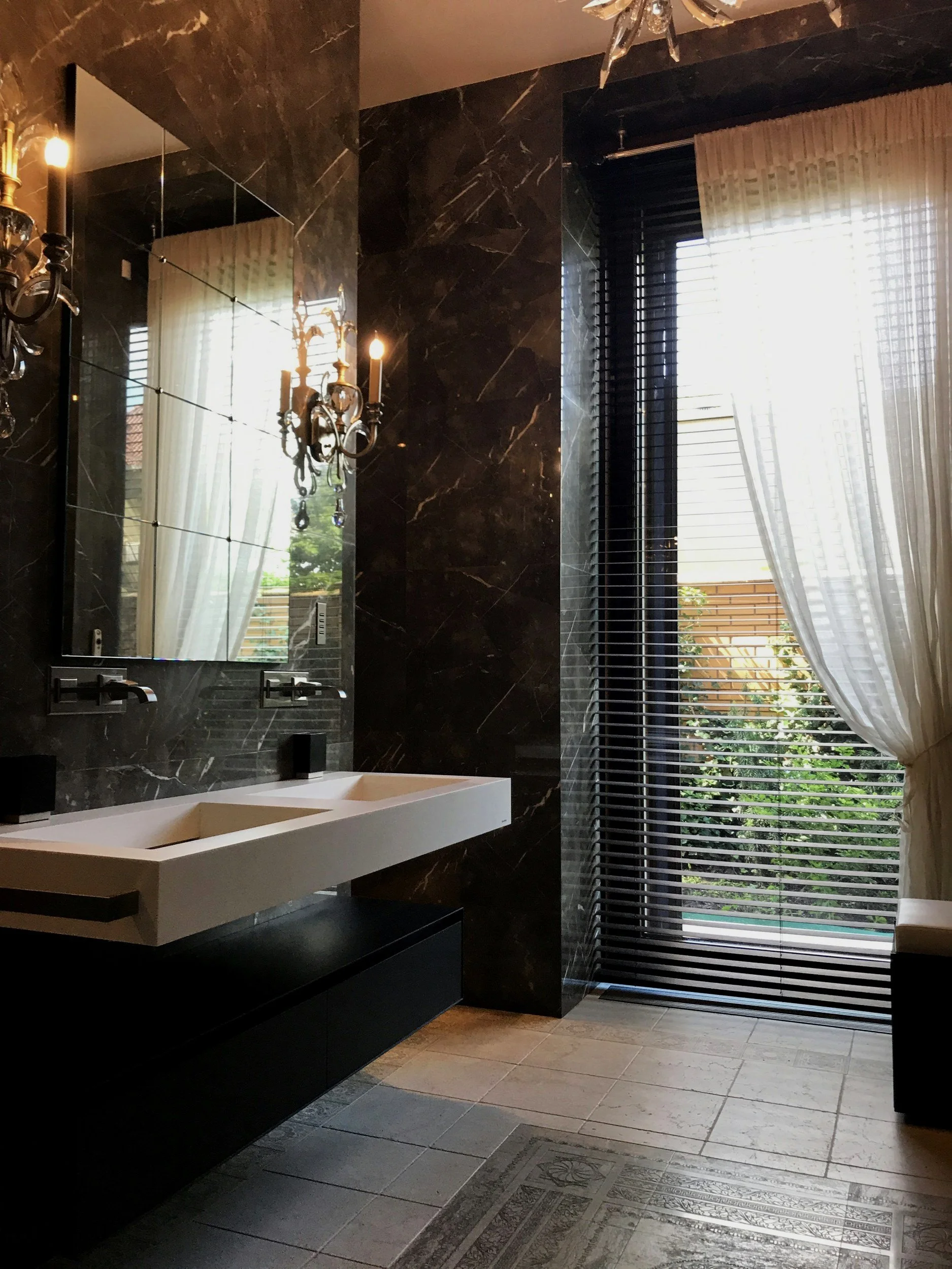 Modern bathroom with black marble walls, a large rectangular white sink, a tall mirror, and a window with blinds and curtains. There is a chandelier and a wall sconce light, and a patterned tile floor.