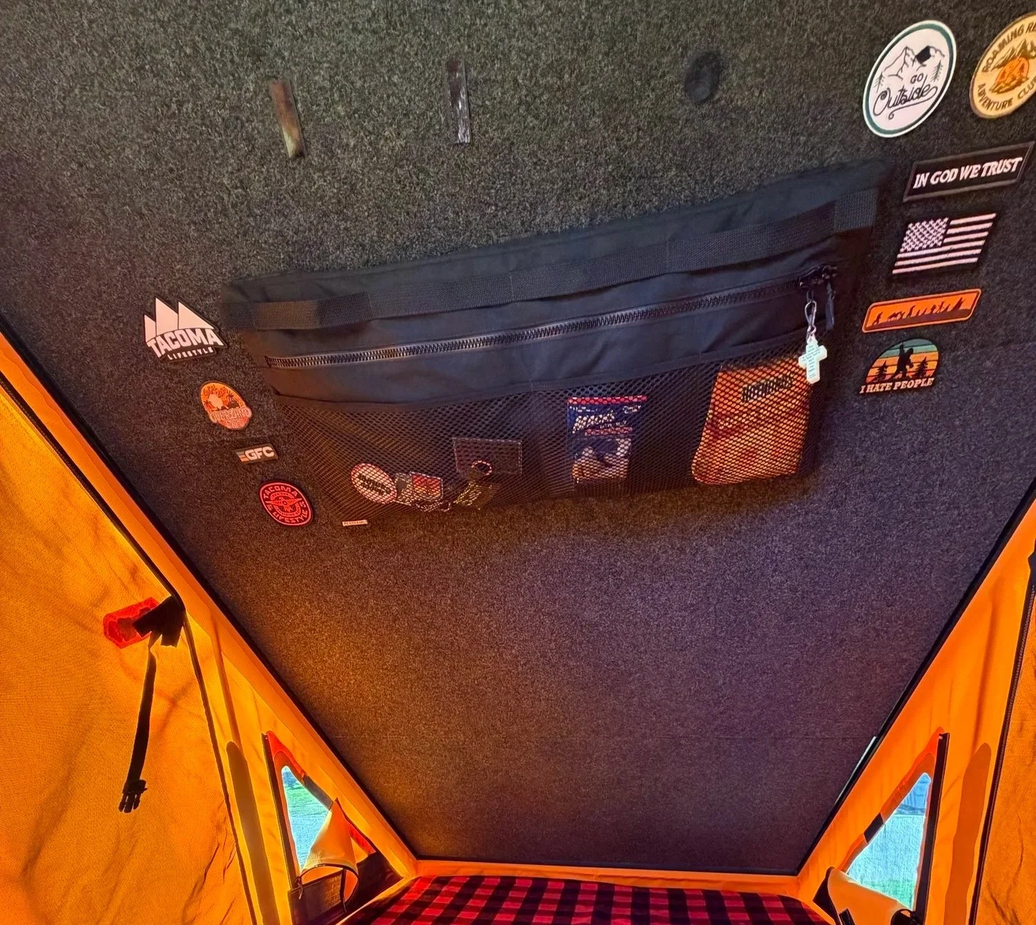 The image shows the interior of a tent with a black ceiling and yellow walls. There is a black mesh storage pocket attached to the ceiling, containing various small items. Several stickers are adhered to the ceiling, including outdoor-themed and patriotic designs. The tent has orange fabric with black straps and a red and black checkered floor.