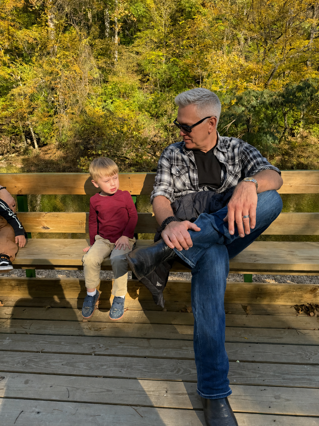A man with gray hair, wearing sunglasses, a black t-shirt, a plaid shirt, and jeans, is sitting on a wooden bench outdoors. There are two young boys nearby, one wearing a maroon shirt and beige pants, and the other partially visible. The background consists of a green and yellow autumn forest.
