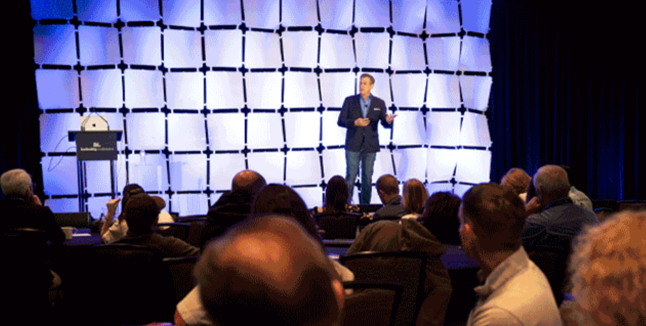 A man in a suit giving a presentation on a stage with a blue grid backdrop. Audience members are seated and listening.