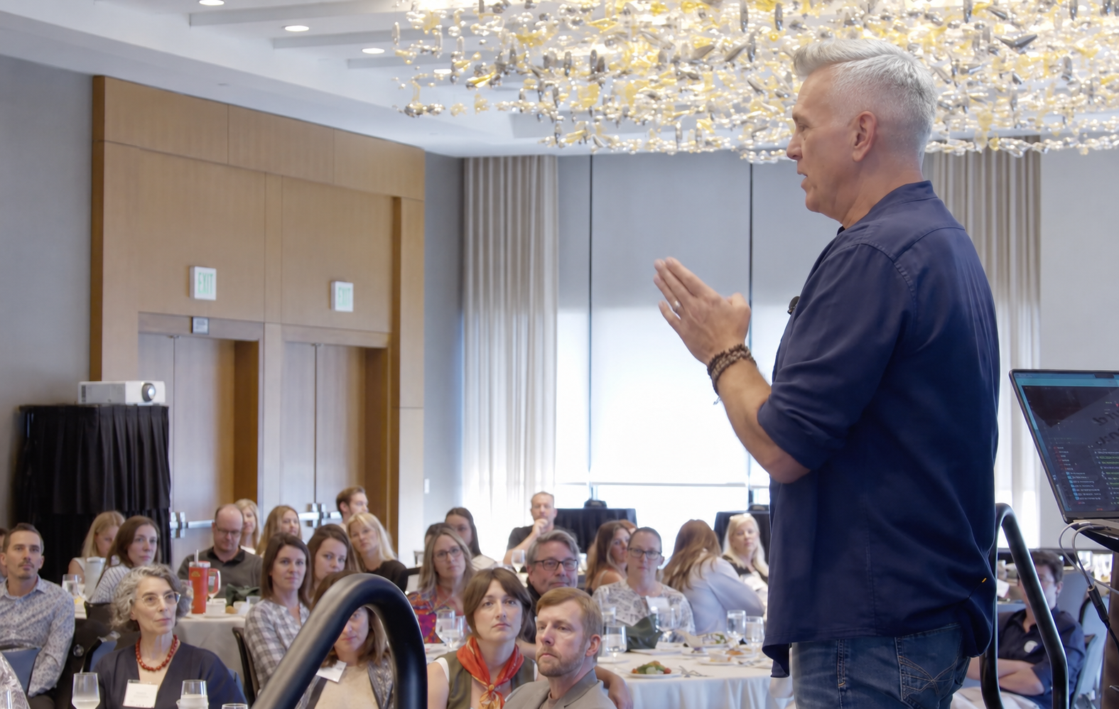 A man giving a presentation to a seated audience in a conference room with a chandelier overhead.