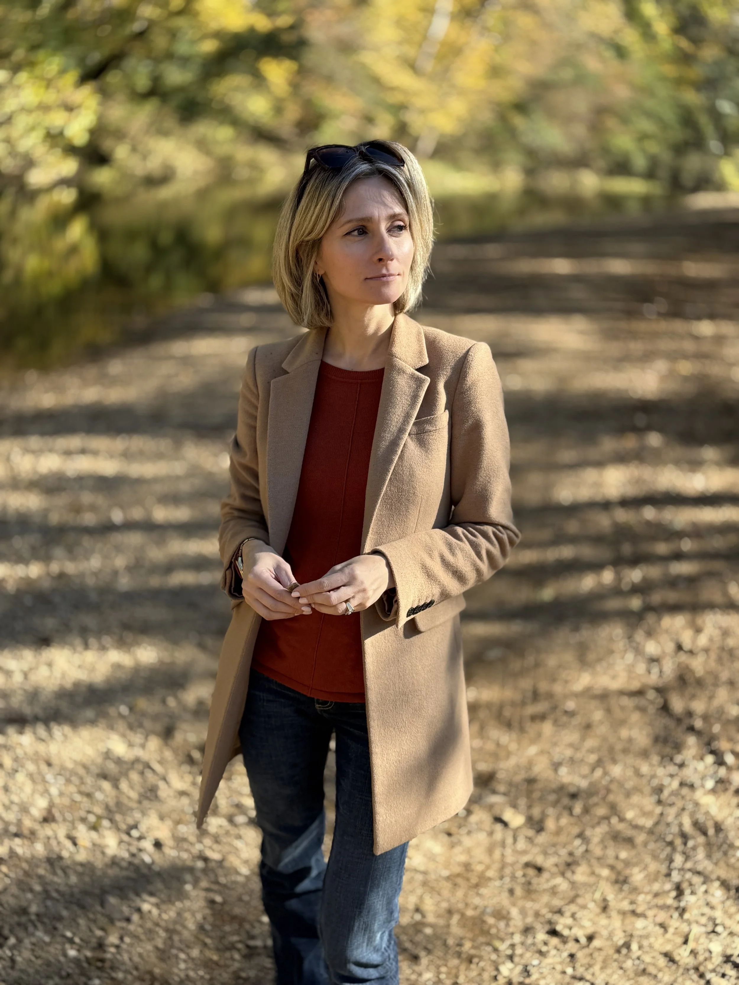 A woman with shoulder-length blonde hair wearing a tan coat, red top, and jeans, standing outdoors on a wooded path in autumn, with some sunglasses on her head and a slight smile.