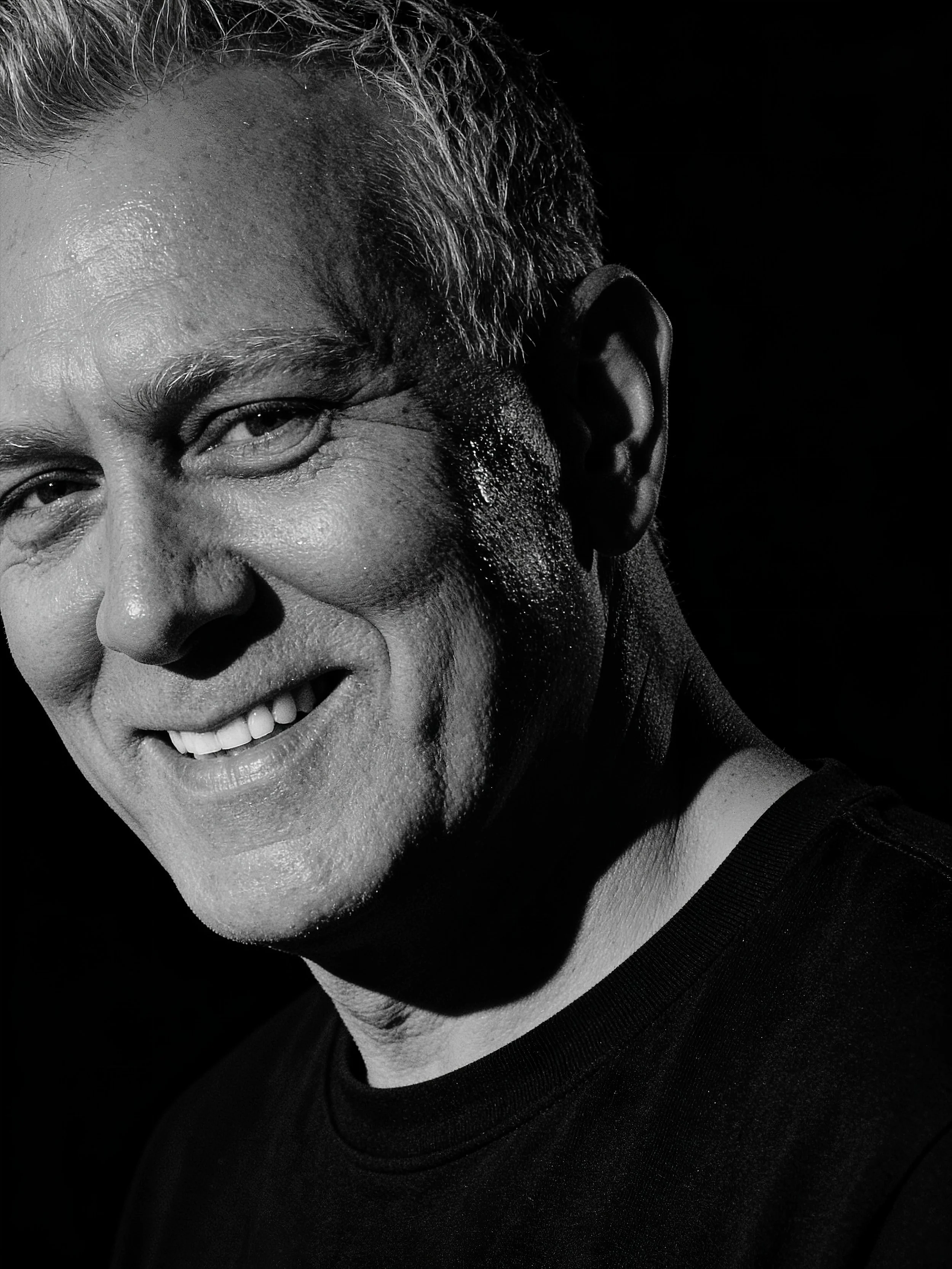 Close-up black and white photo of a smiling person, showing part of their face, ear, and neck, with textured skin and short hair, against a dark background.
