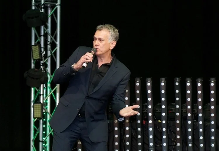 Man in a dark suit holding a microphone on a stage with lighting equipment behind him.