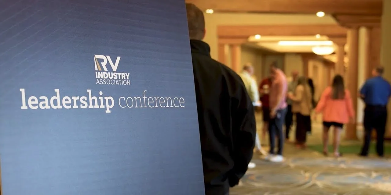 A man standing next to a blue sign for the RV Industry Association leadership conference, with a group of people in the background in a conference center hallway.