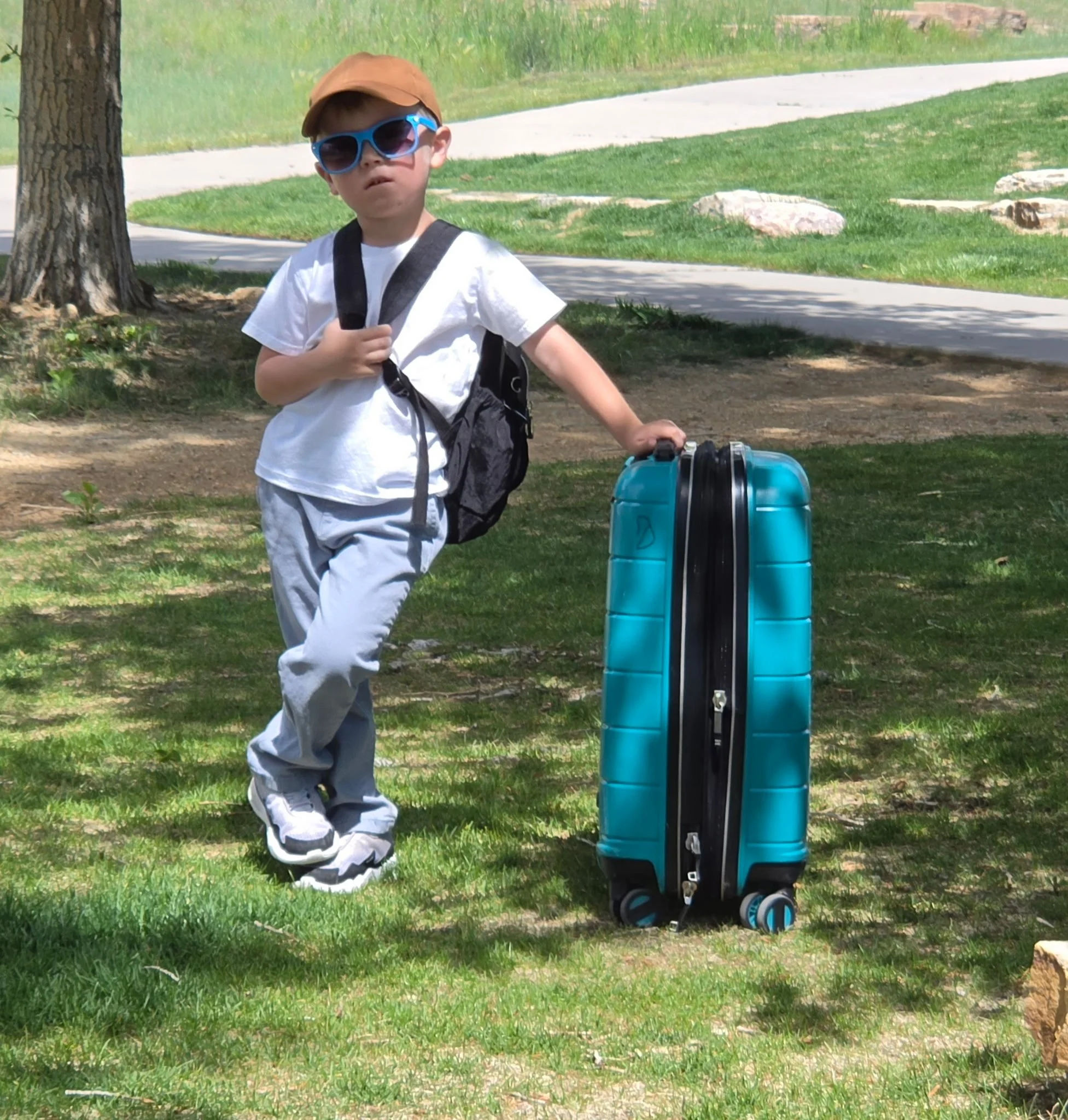 A young boy wearing sunglasses, a brown baseball cap, a white t-shirt, and gray pants, standing outdoors beside a teal suitcase with wheels. He has a black backpack on his shoulders, and he is holding onto his backpack strap with one hand and the handle of the suitcase with the other. The background includes a grassy area with a tree, rocks, a sidewalk, and a blurred patch of bushes.