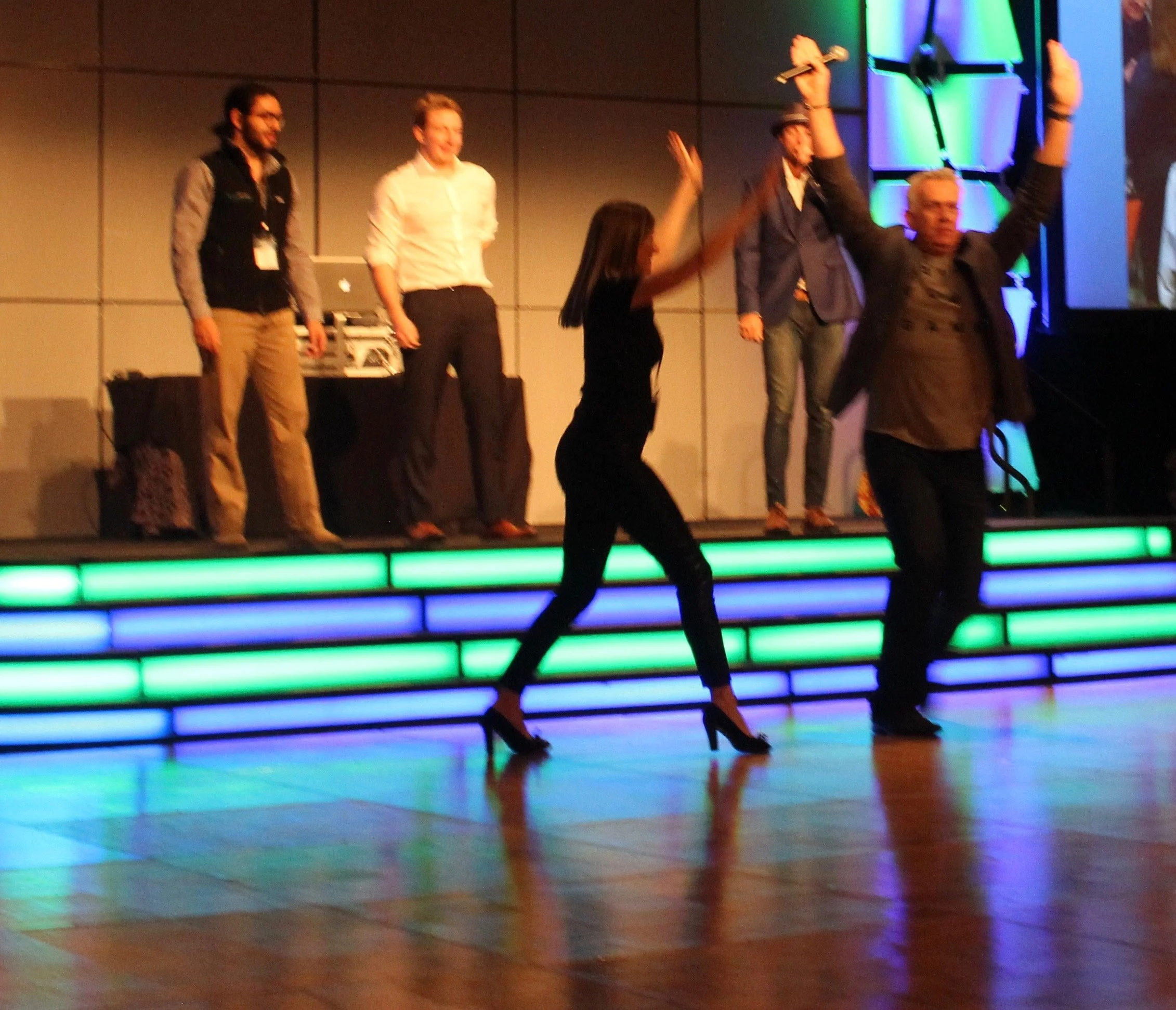 People dancing on a stage with colorful illuminated steps, one person with long hair appears to be performing a dance.