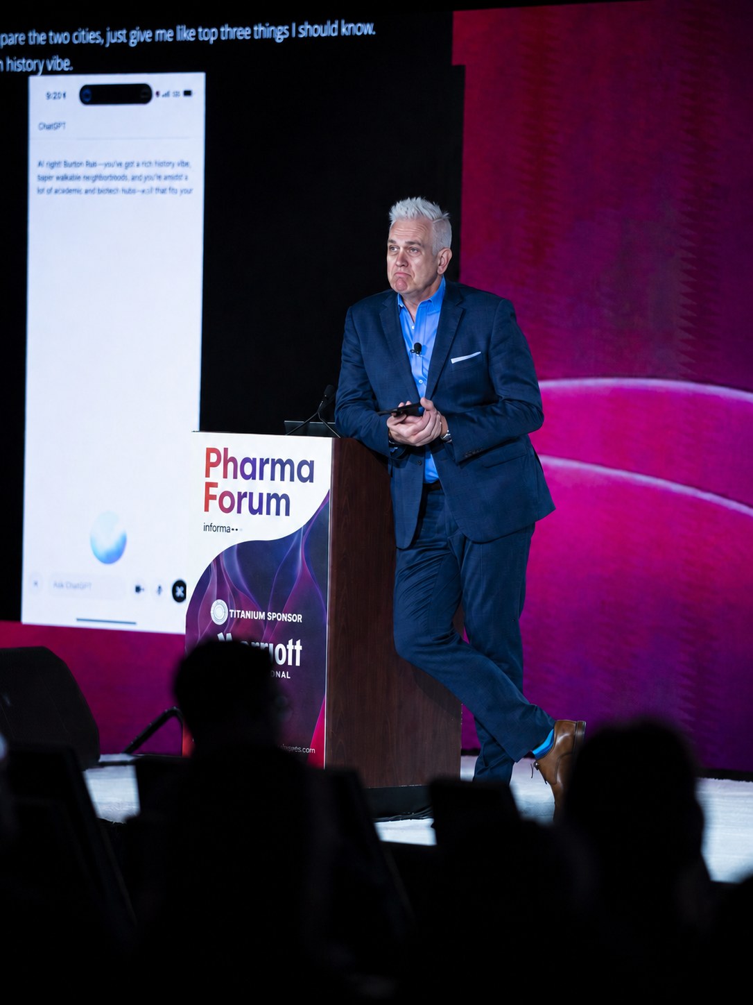 A man in a blue suit with gray hair stands on stage at Pharma Forum, holding a phone, with a large screen displaying a phone screen and a purple background behind him.