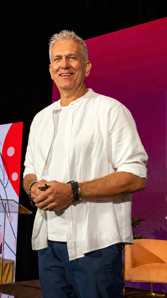 Smiling man with gray hair wearing a white shirt and dark pants standing on stage at an event with a colorful background.