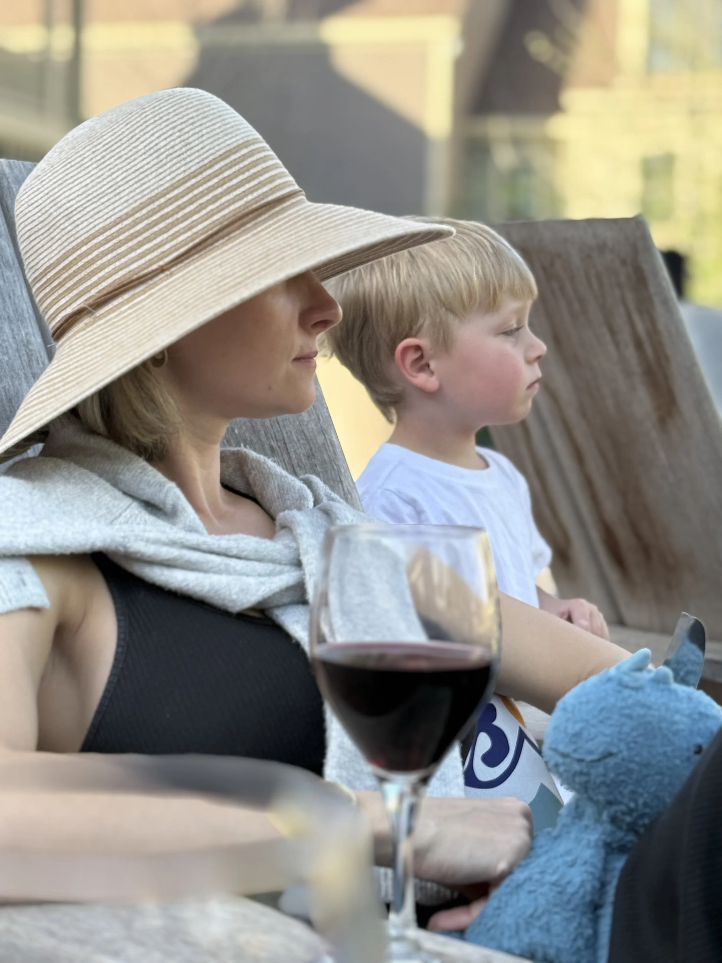 A woman wearing a large wide-brimmed sun hat, sitting outdoors with a young boy. They are seated at a table with a glass of red wine in the foreground and are facing sideways with their eyes closed or looking down. They appear relaxed, enjoying a peaceful moment in a sunny outdoor setting.