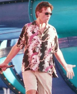 Person wearing sunglasses and a tropical shirt walking near a large slide at an amusement park.