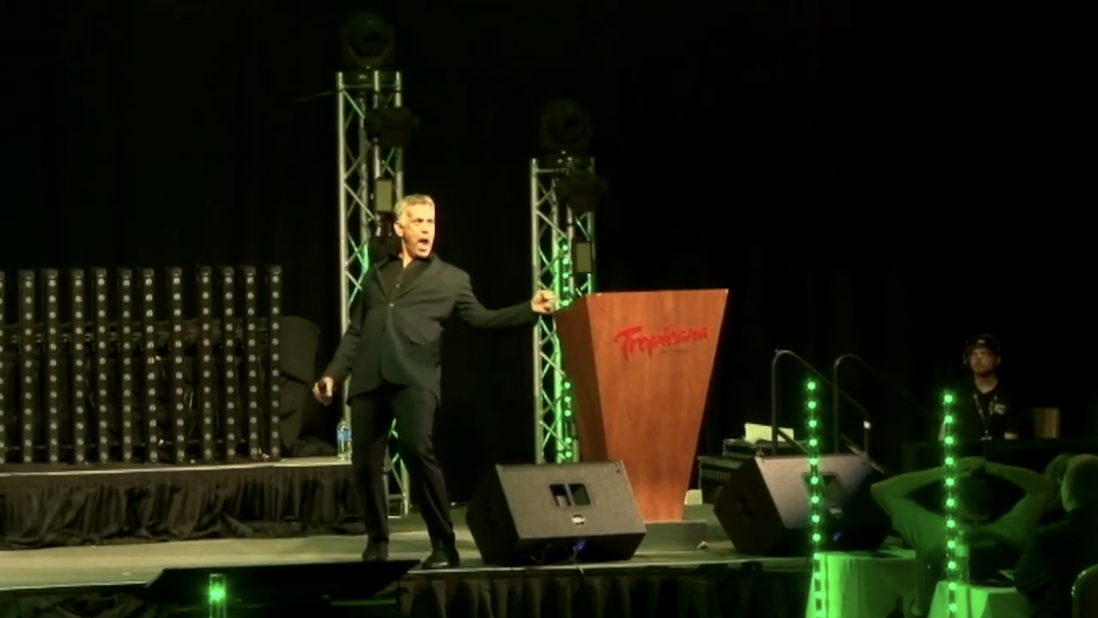 Man wearing black suit and black shirt speaking on stage at conference with a wooden podium labeled 'Tropicana' and large black curtain backdrop, with stage lighting and green decorative lights.