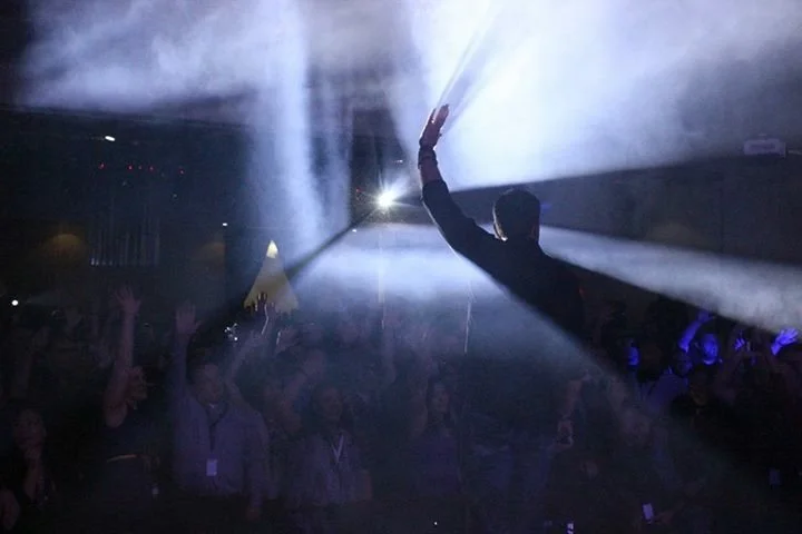 A performer on stage with one arm raised and facing a crowd at a concert or event, with dramatic lighting and smoke effects.