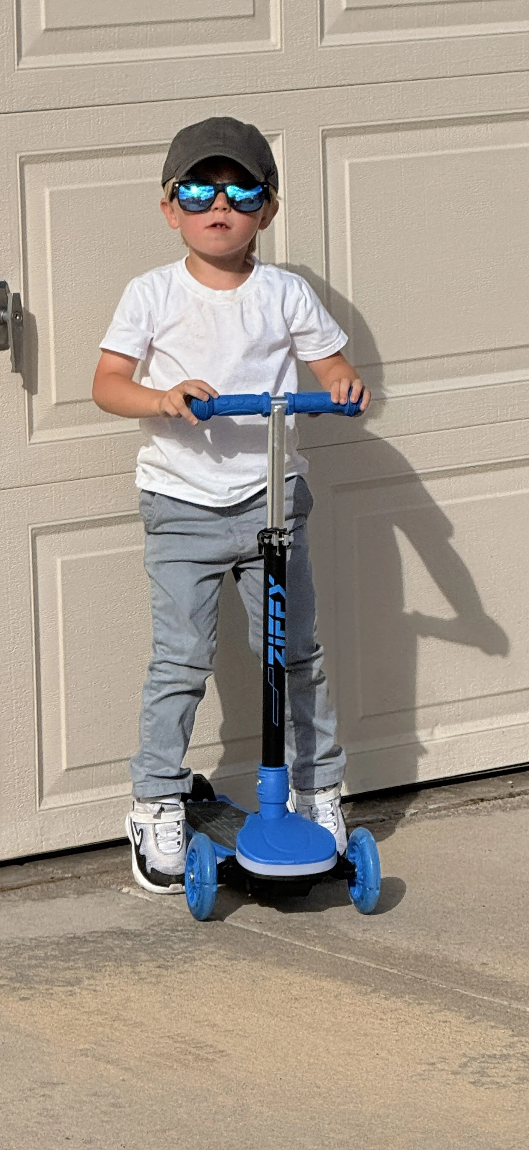 A young boy wearing a gray cap, sunglasses, a white t-shirt, gray pants, and sneakers, standing on a blue and black scooter in front of a beige garage door.